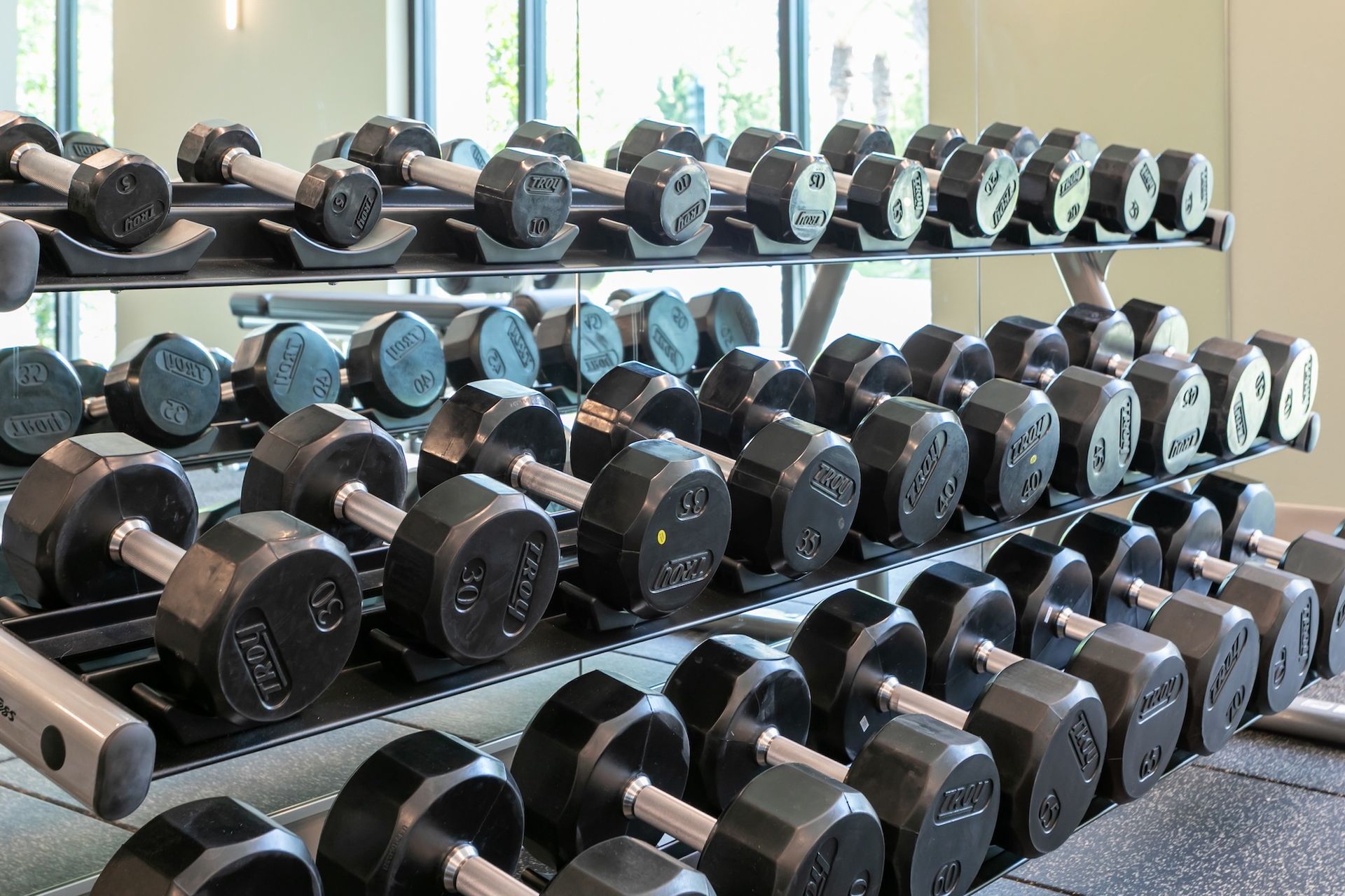 Dumbbells on a rack in a gym setting. Weights are black with chrome handles, arranged neatly on multiple tiers.