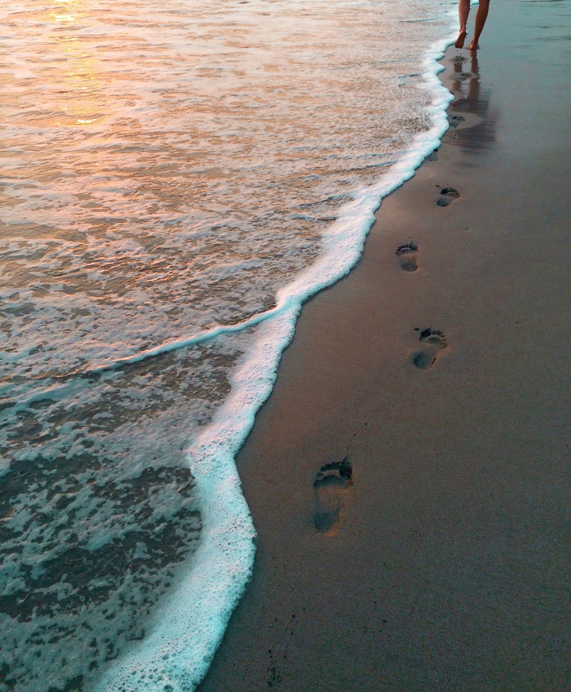Footprints in wet sand as waves crash, person walking towards sunset.