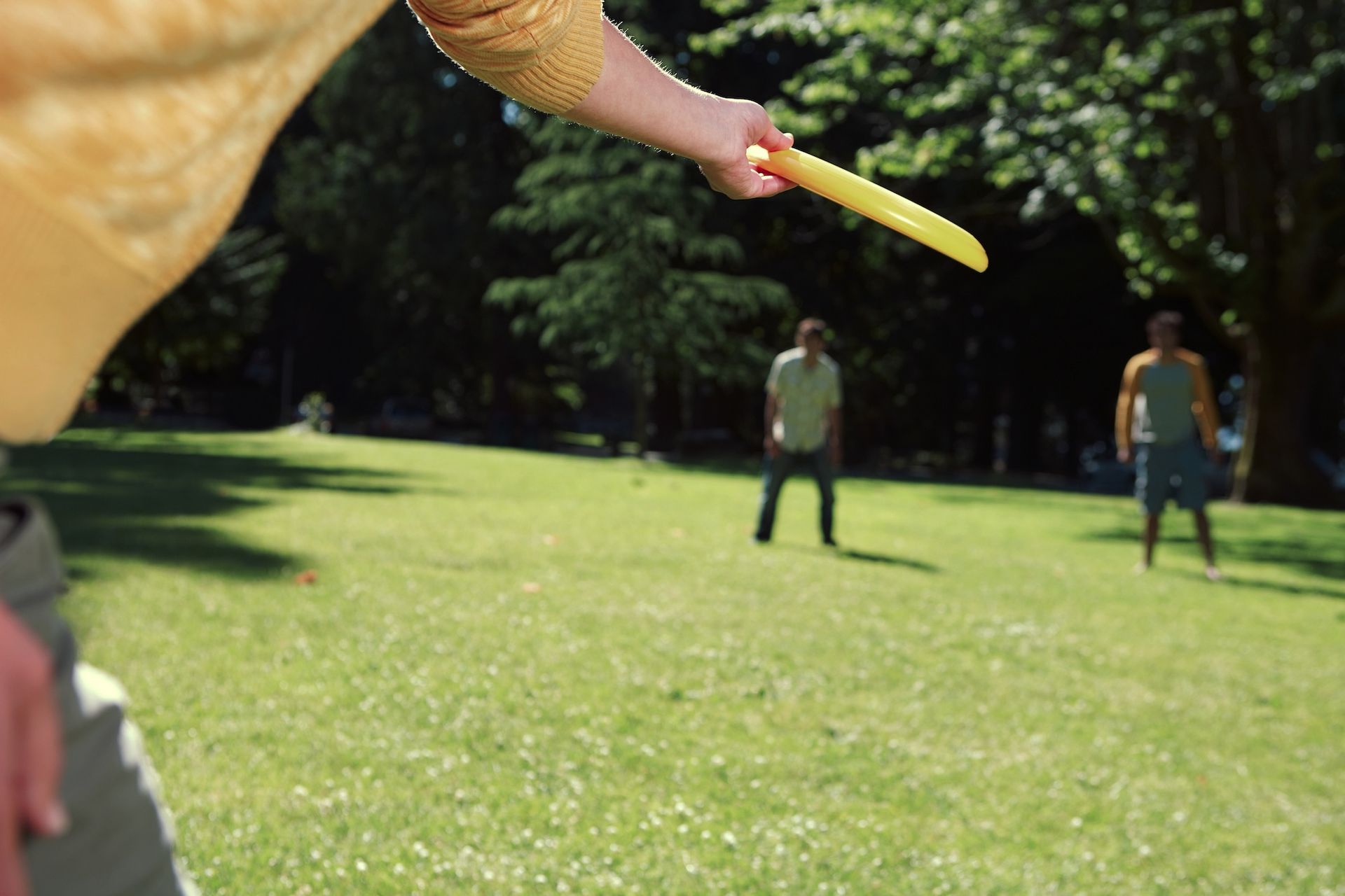 Person throwing a yellow frisbee in a sunny park; two people stand ready to catch it.
