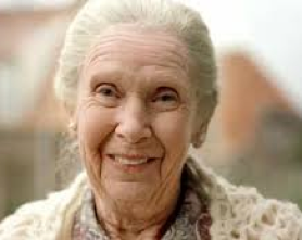 Smiling elderly woman with white hair in a light patterned sweater, outdoors with blurred houses behind her