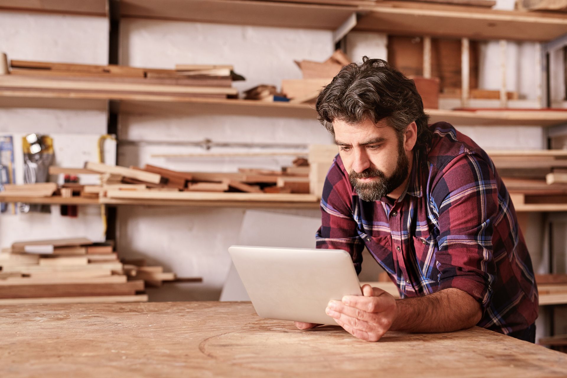 Man in plaid shirt using a tablet in a wood workshop, leaning on a wooden table. Shelves with wood in the background.