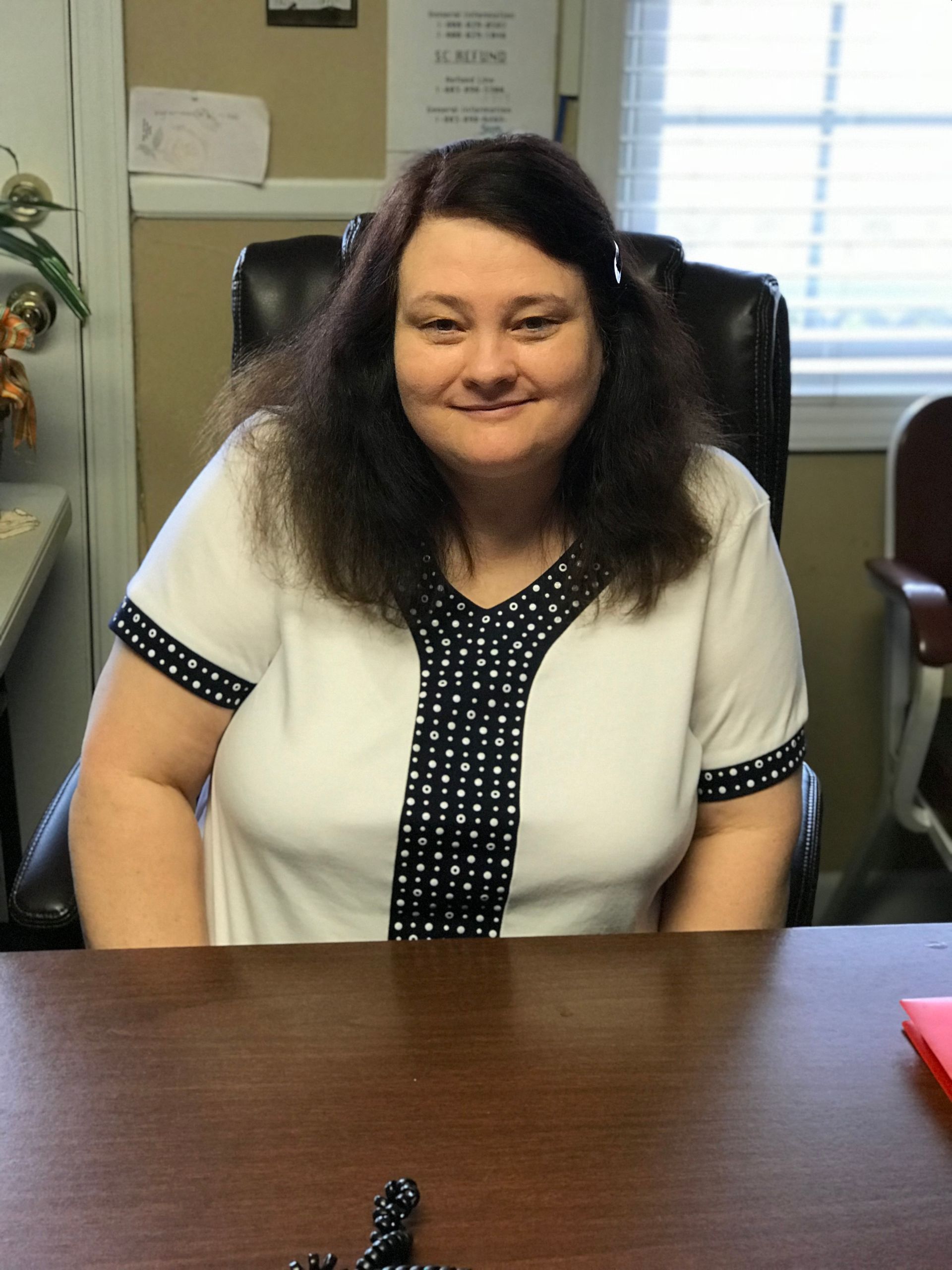 A woman is sitting at a desk in front of a window and smiling