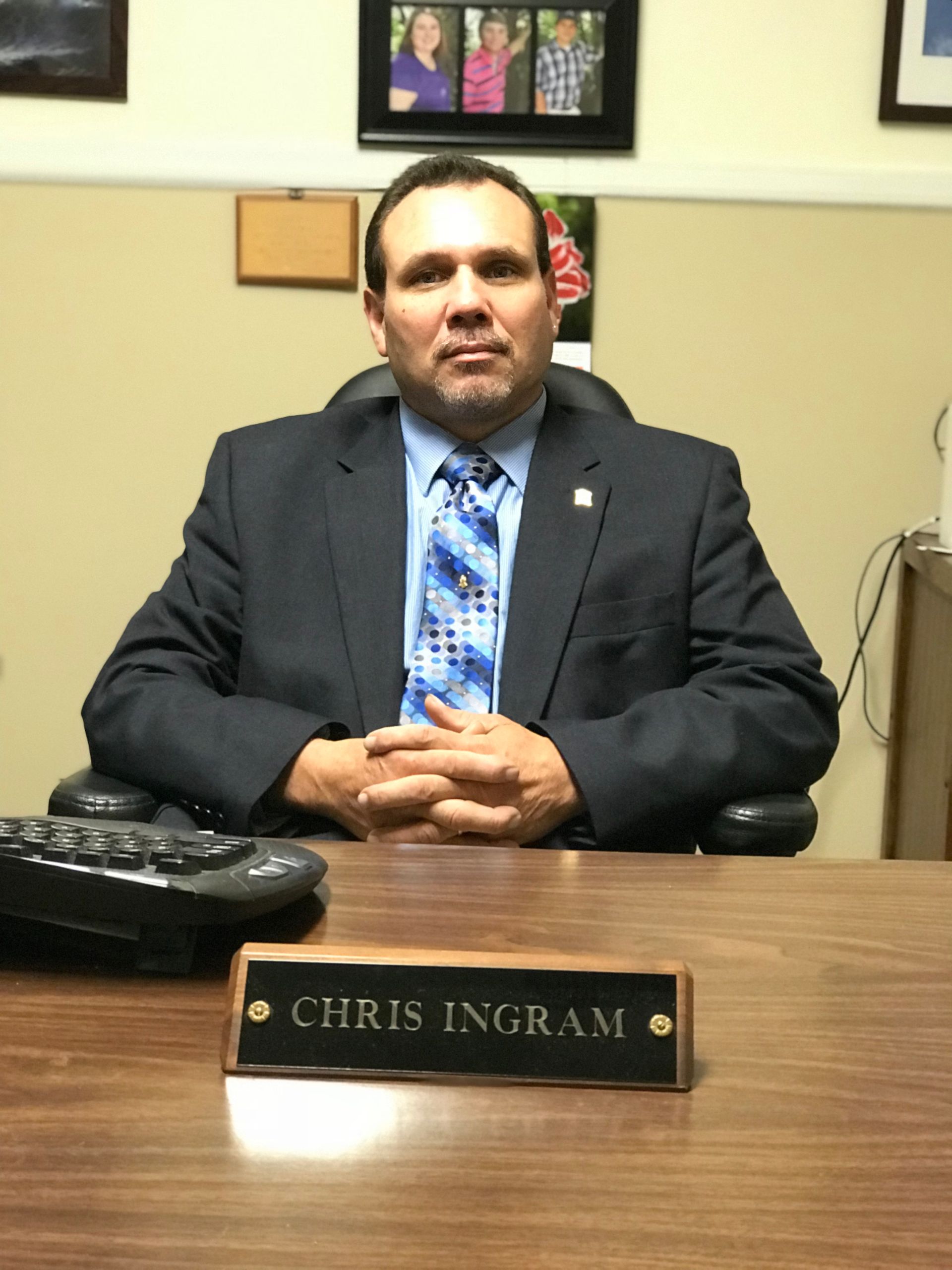 A man in a suit and tie sits at a desk with a name plate that says chris ingram