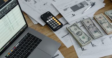 A laptop computer is sitting on a wooden table next to a pile of money and a calculator.