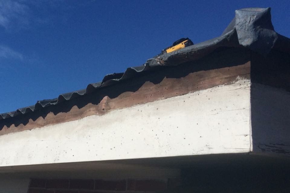 Close-up view of a building's roof edge, showing a white concrete structure with a corrugated, dark-colored roofing material against a blue sky.