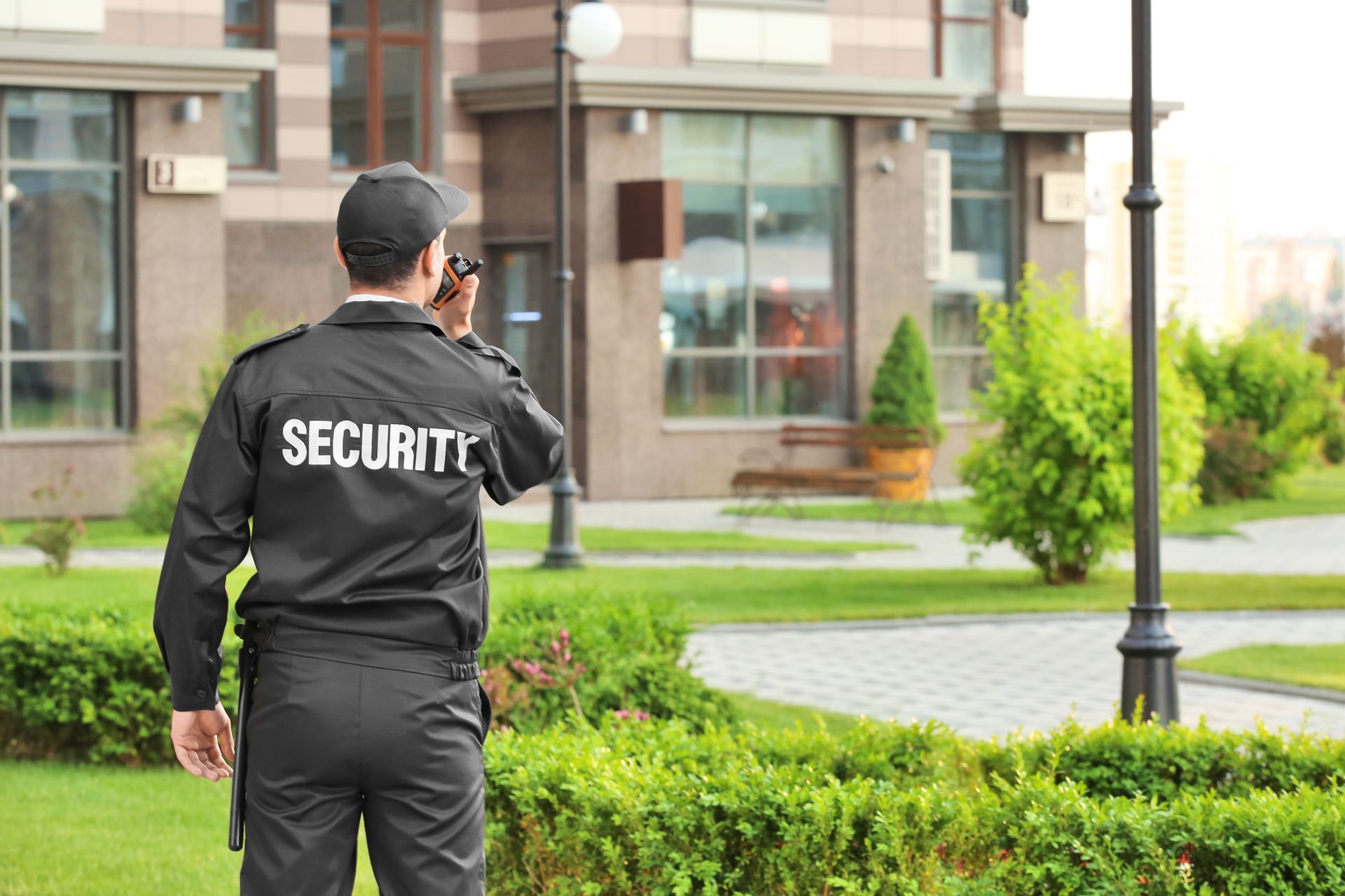 Security guard in black uniform standing in a courtyard near a building and lamp post.