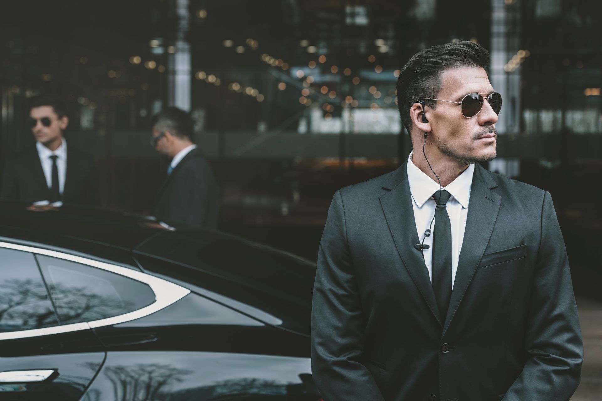 Man in dark suit and sunglasses standing beside a black car outside a modern building at night.