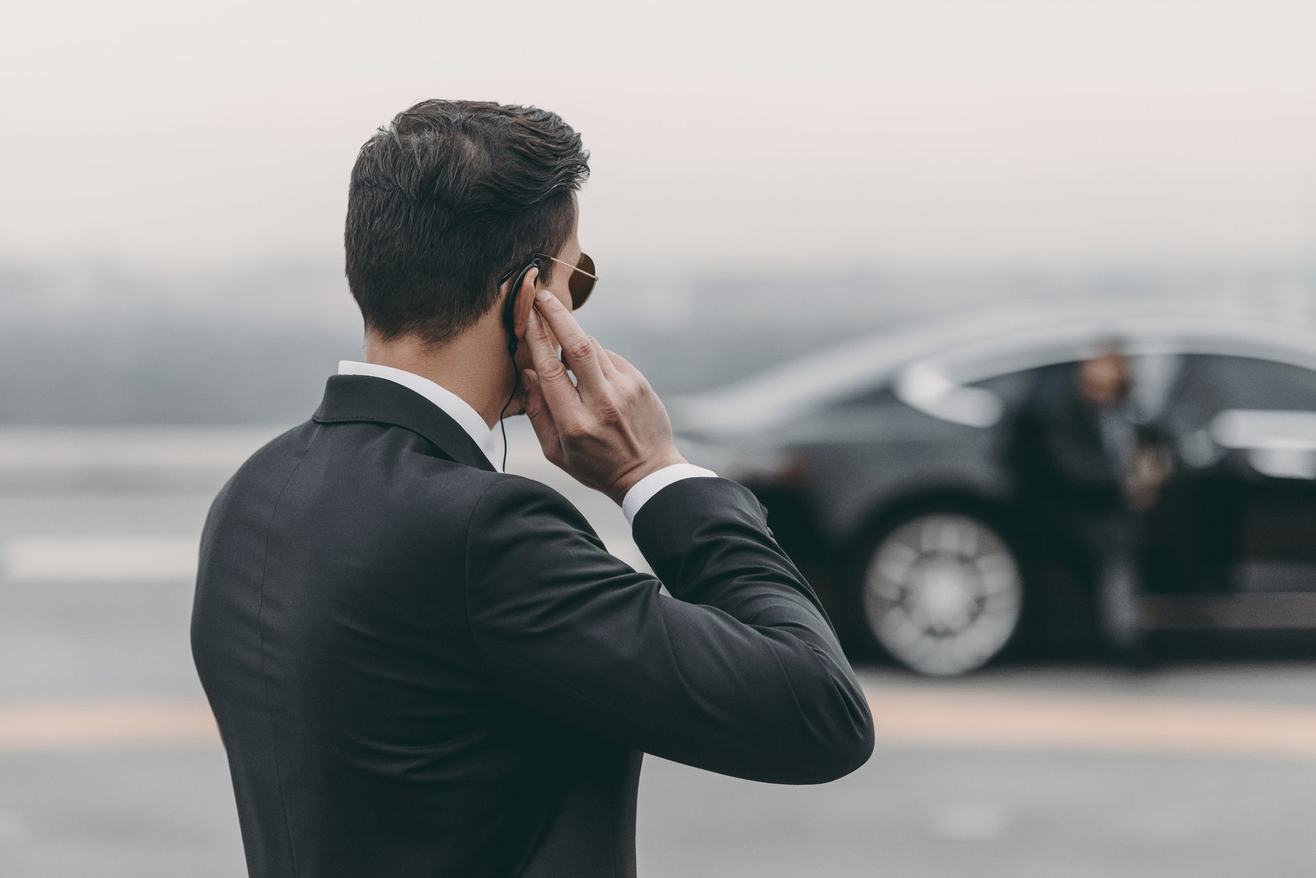 Man in a black suit speaking on a phone beside a black luxury car on a roadside.