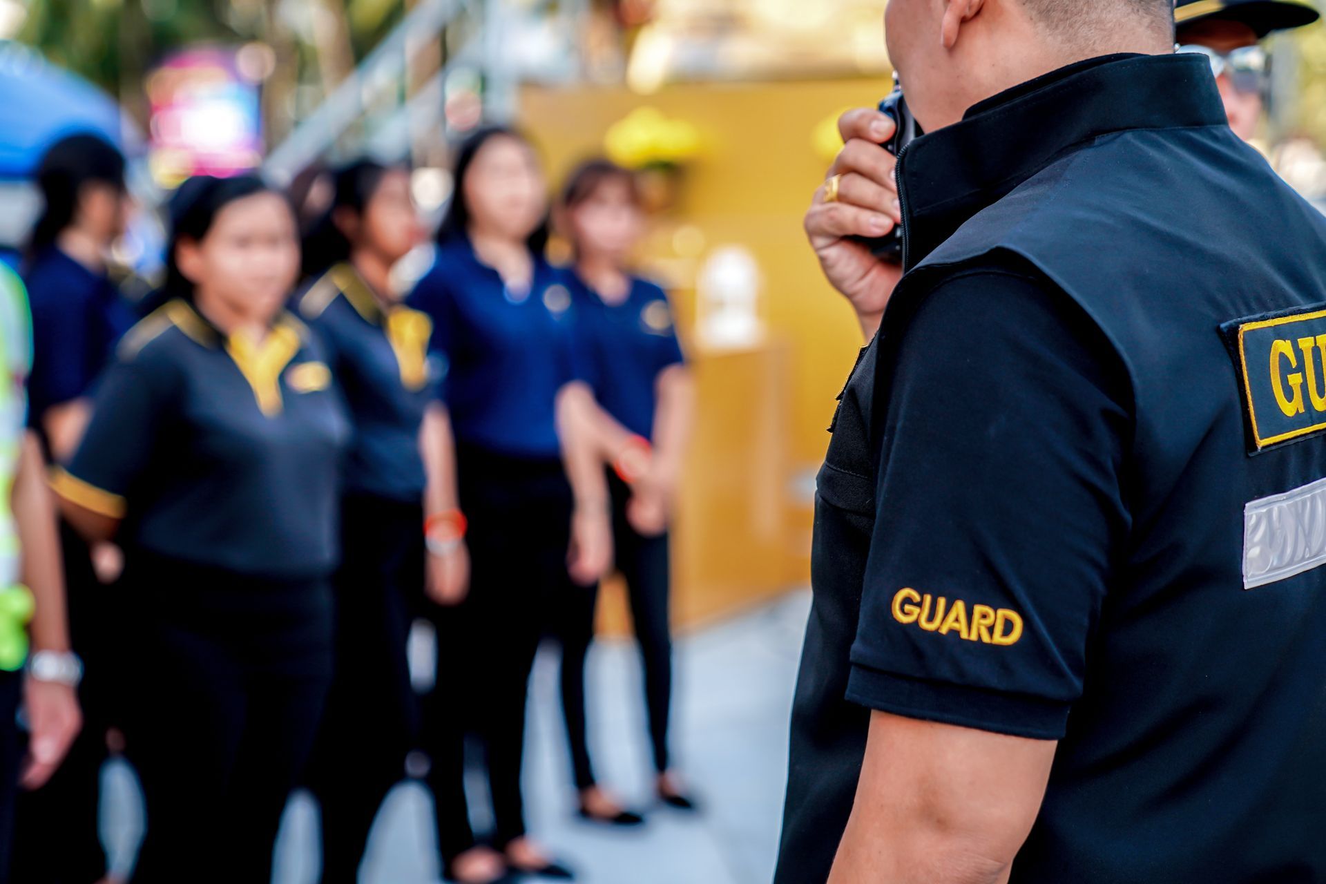 Security guard in black shirt speaking to a line of people in blue uniforms outdoors.