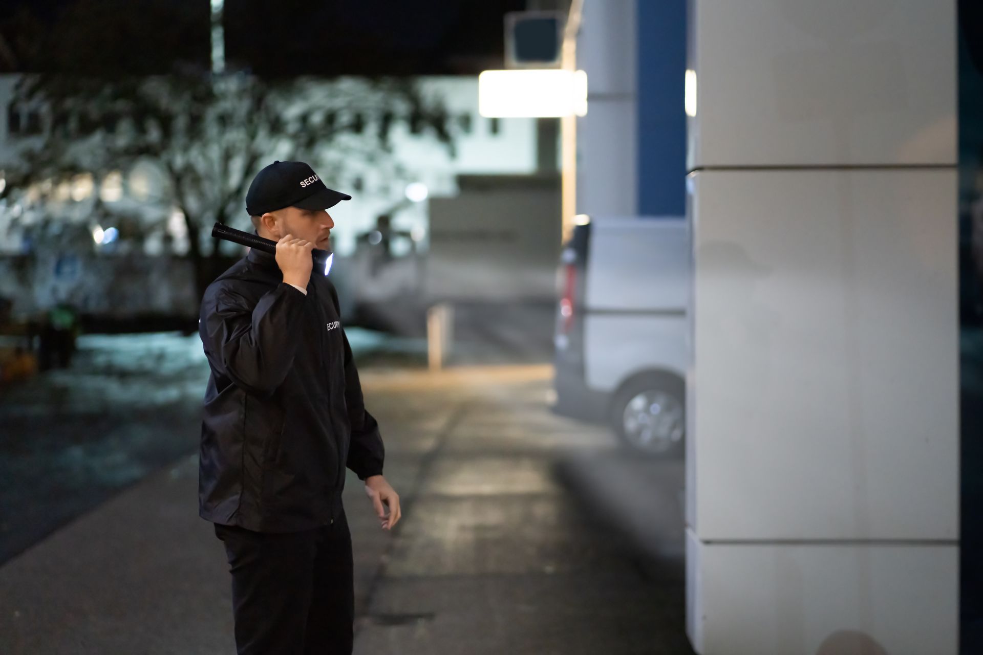 Person in a black cap and jacket stands outdoors beside a building at night.