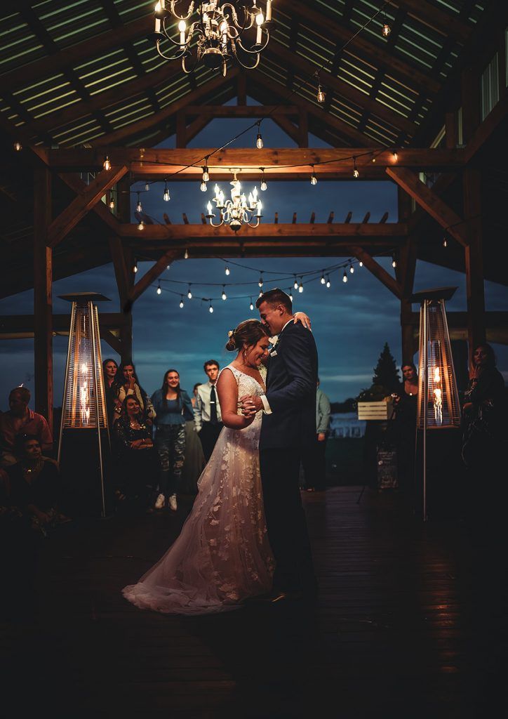 Couple dances at a wedding reception in a wooden barn, under string lights.