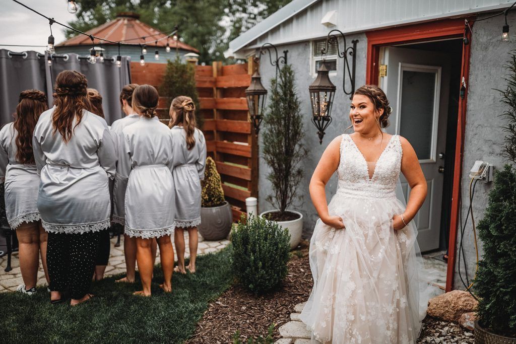Bride in wedding dress, surprised expression, stands outside a building. Bridesmaids in robes stand nearby.