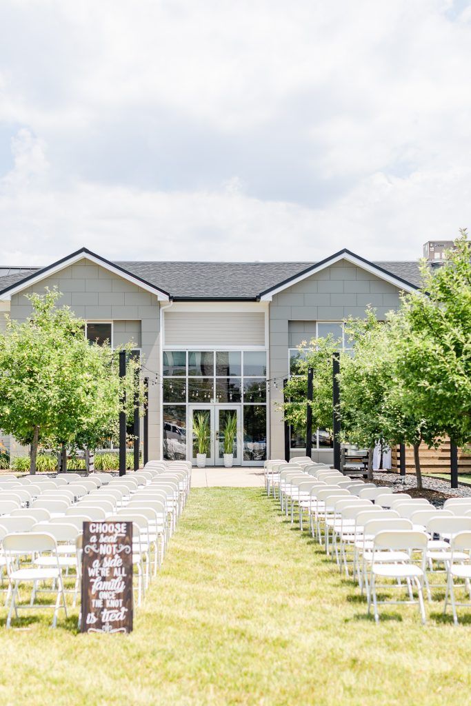 Wedding ceremony setup: white chairs line a grassy aisle leading to a modern building with glass doors, trees flank sides.