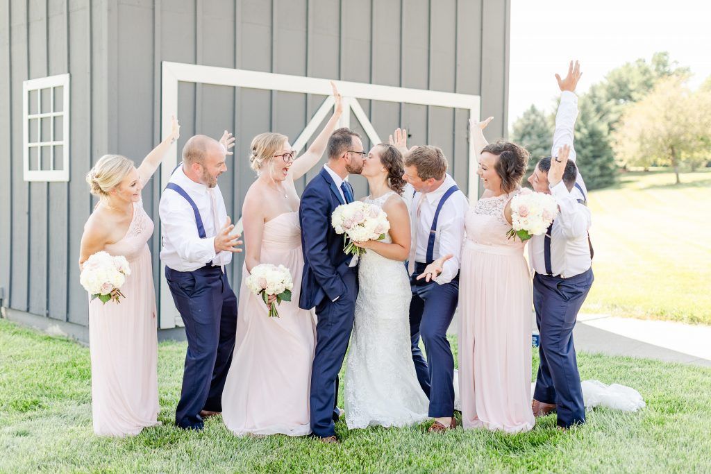 Wedding party celebration bride and groom kiss, surrounded by bridesmaids in pink and groomsmen in navy in front of a barn.