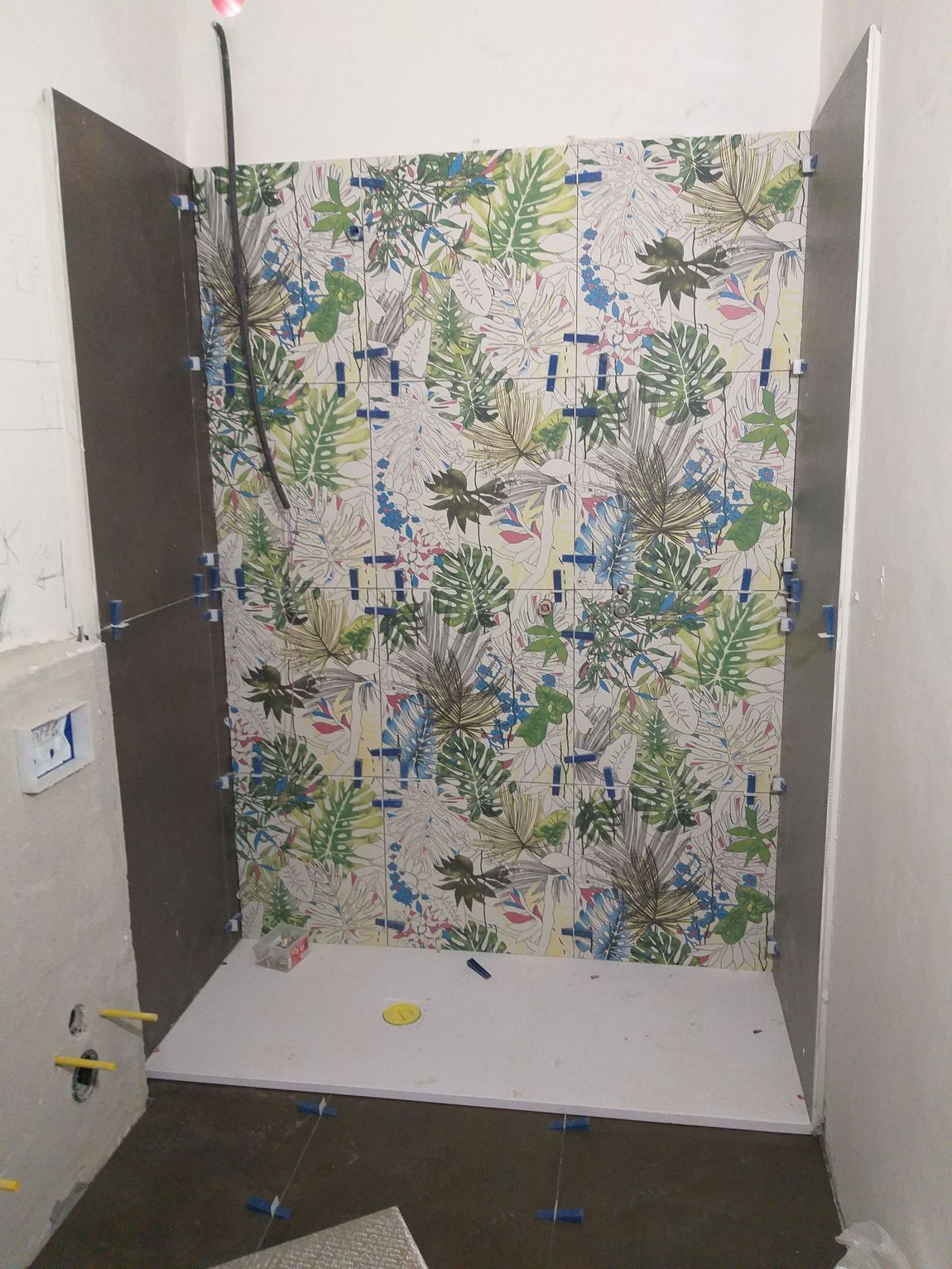 Piccolo bagno con carta da parati a motivi floreali, porte scure e pavimento bianco in fase di ristrutturazione.