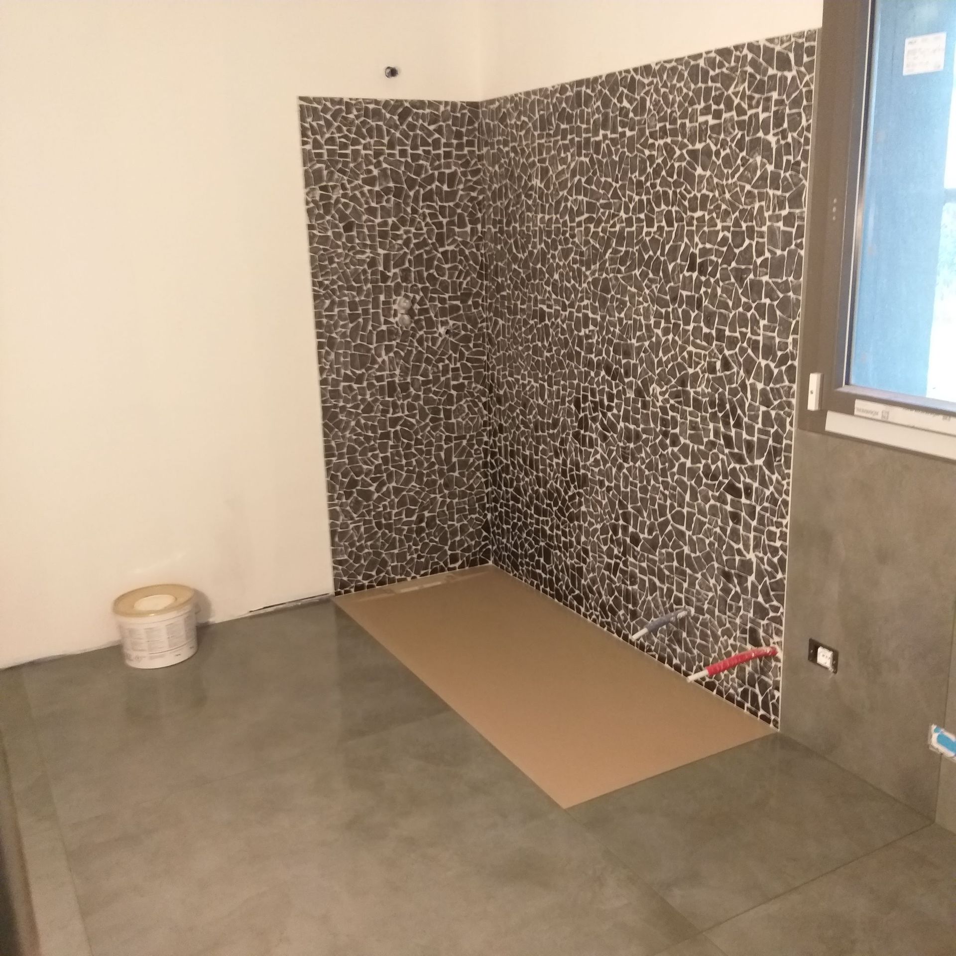 Stanza parzialmente rifinita con una parete decorativa a motivi geometrici, pavimento grigio e cartone sul pavimento vicino a una finestra.