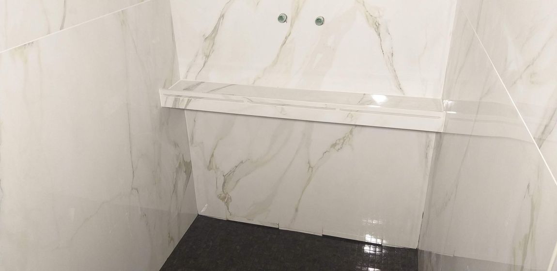 Piccolo angolo doccia piastrellato con mensola bianca e pareti effetto marmo.