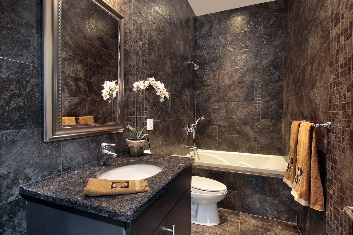 Bagno in marmo scuro con mobiletto, specchio, lavabo, WC, vasca da bagno e asciugamani arancioni.