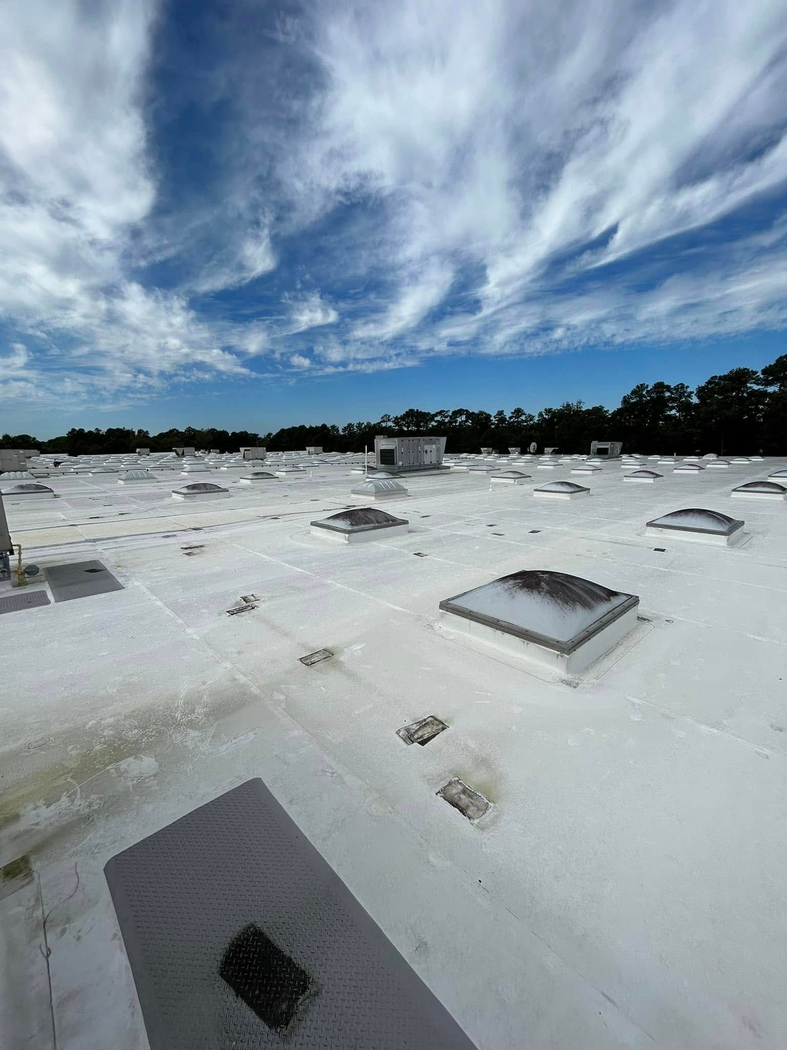 A white, flat commercial roof with multiple skylights under a bright blue sky with wispy clouds.