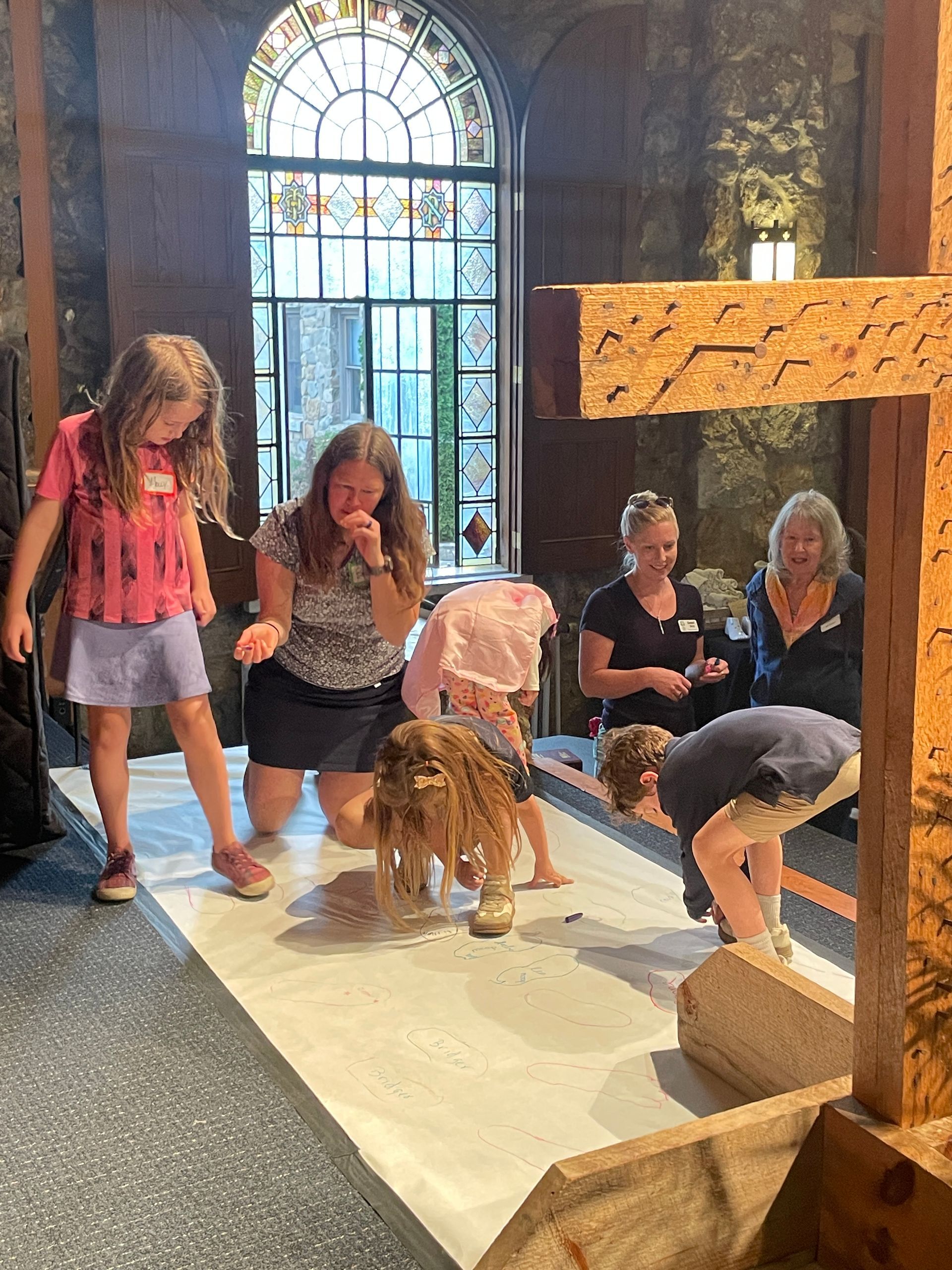 Children and adults tracing feet on white butcher paper with a wooden cross in the foreground.