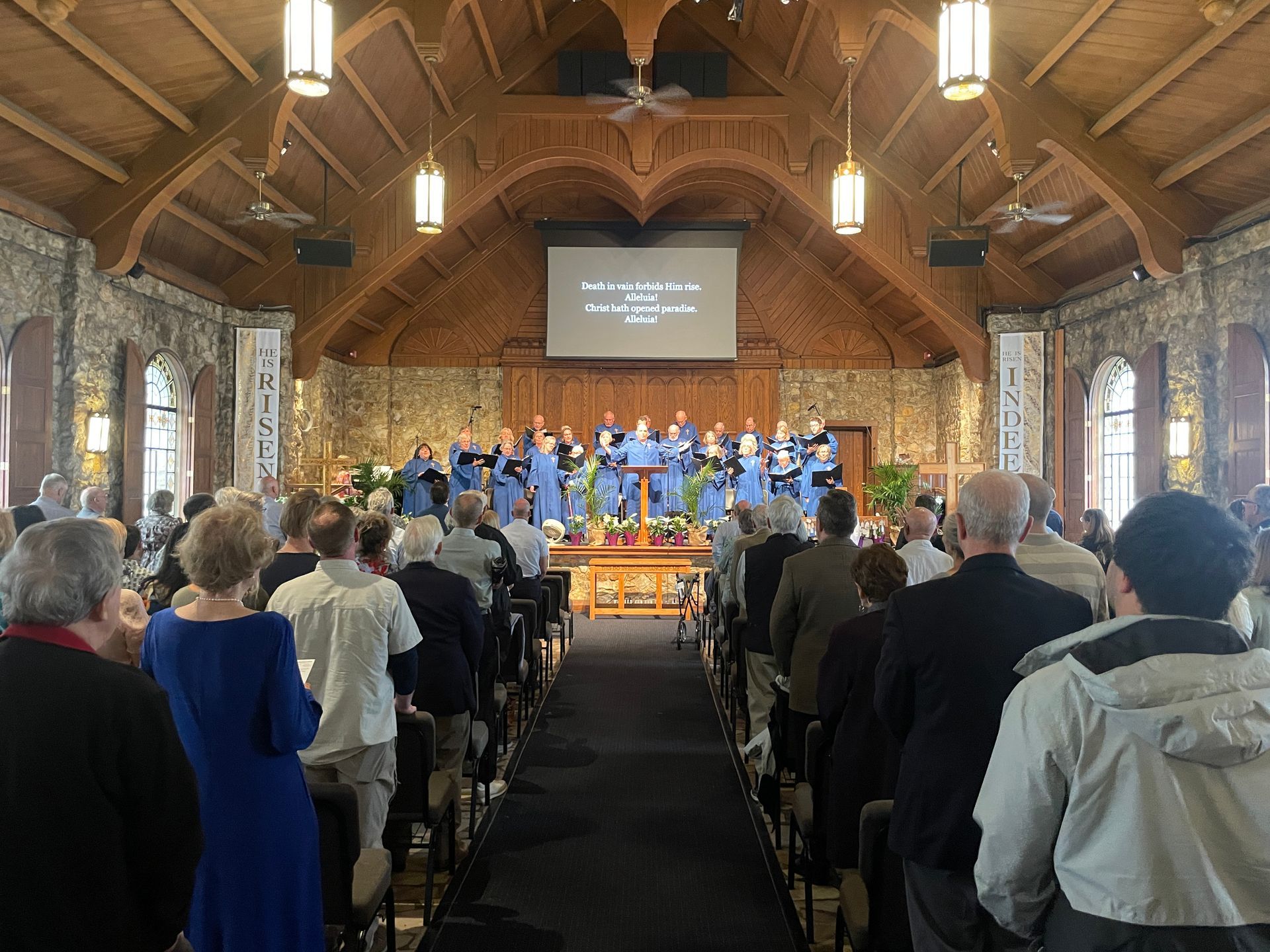 People standing in a stone and wood chapel facing a stage with a blue-clad choir and a projection screen above.