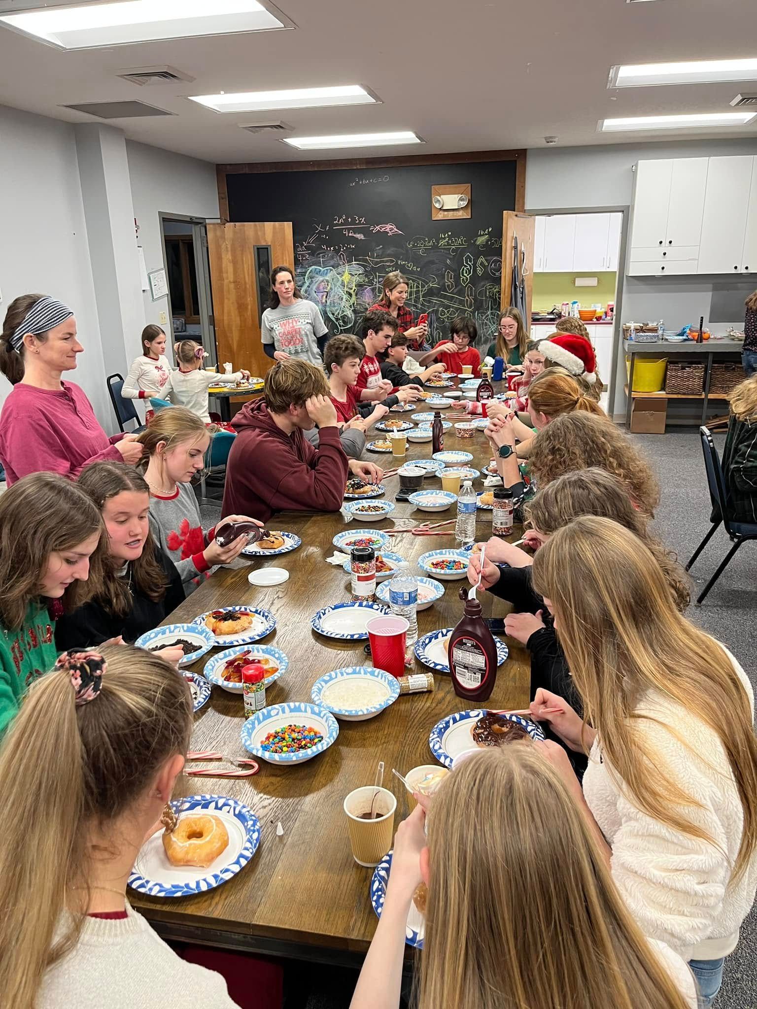Long table full of teenagers decorating doughnuts with a variety of toppings.