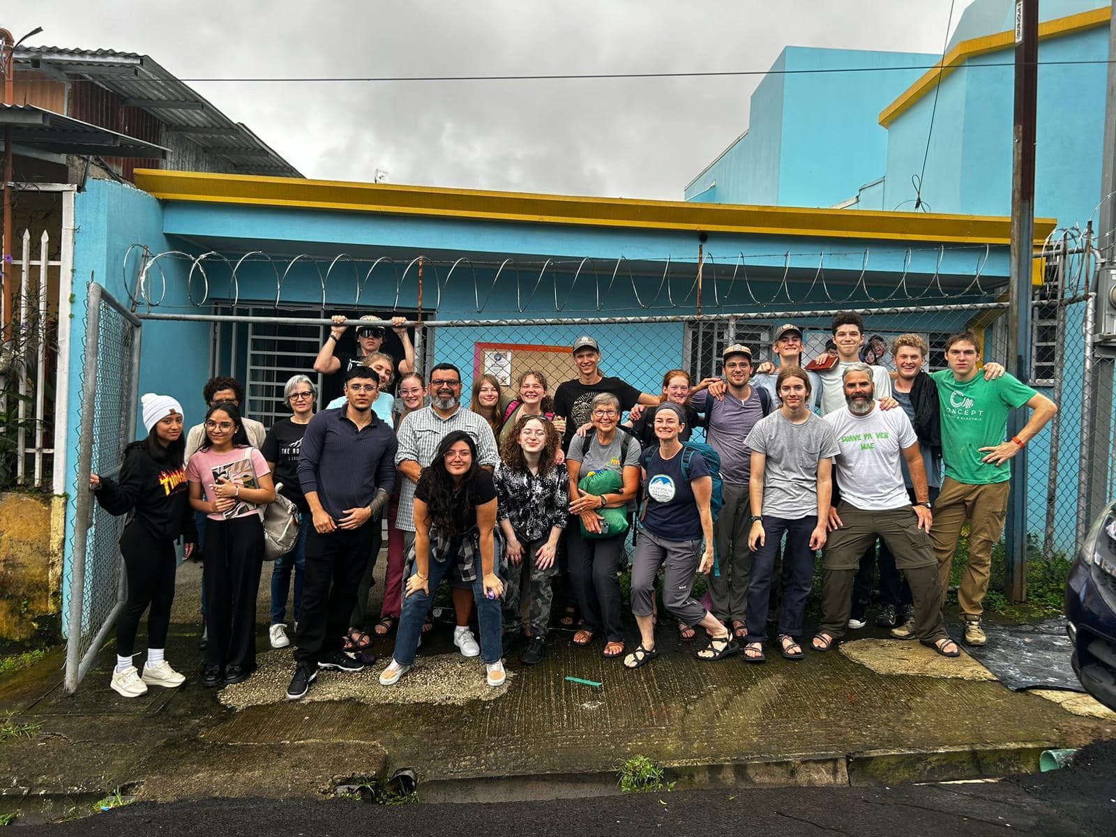 Large group of people standing in front of a blue building fenced in for security.