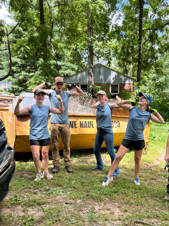 Four teenagers in matching blue shirts show off their muscles in front of a yellow dumpster of trash.