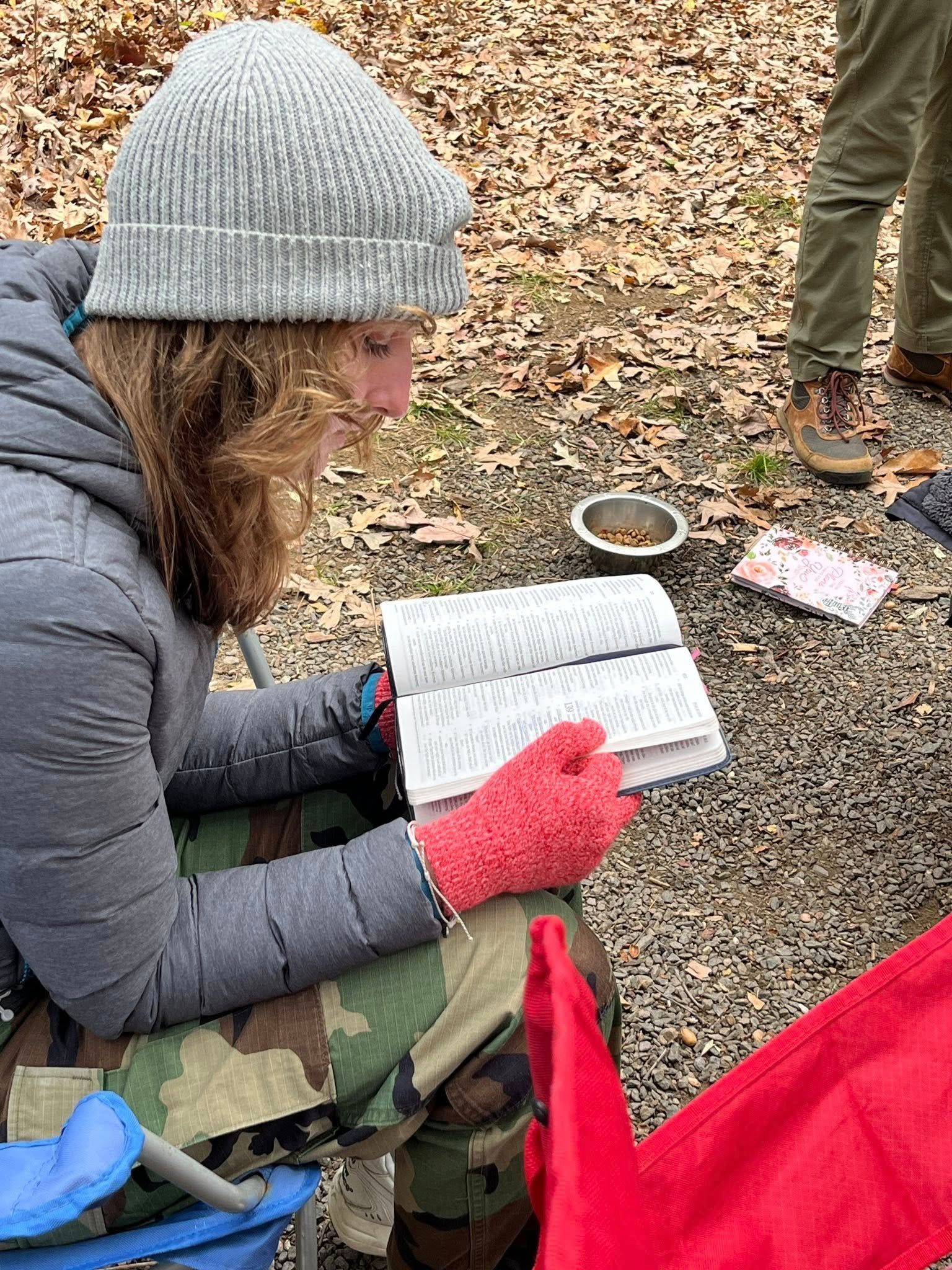 Person in a gray beanie and red gloves reading a Bible outside in a camping chair.