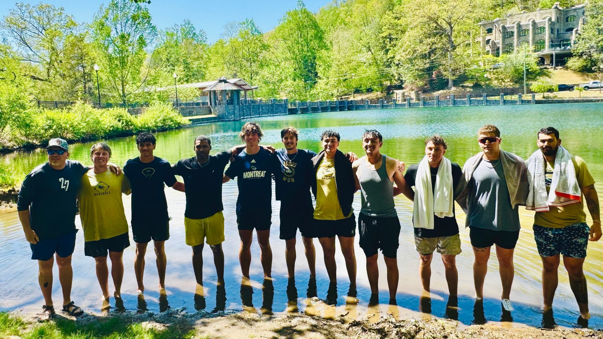 11 men standing in ankle deep water at the edge of a small lake with trees and a stone wall behind them.