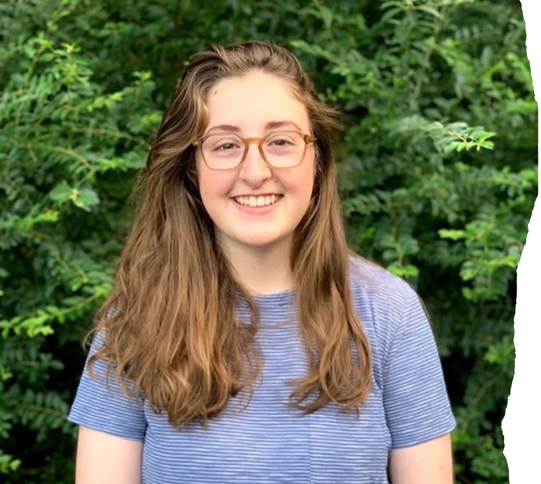Woman with long brown hair and glasses stands in front of greenery.