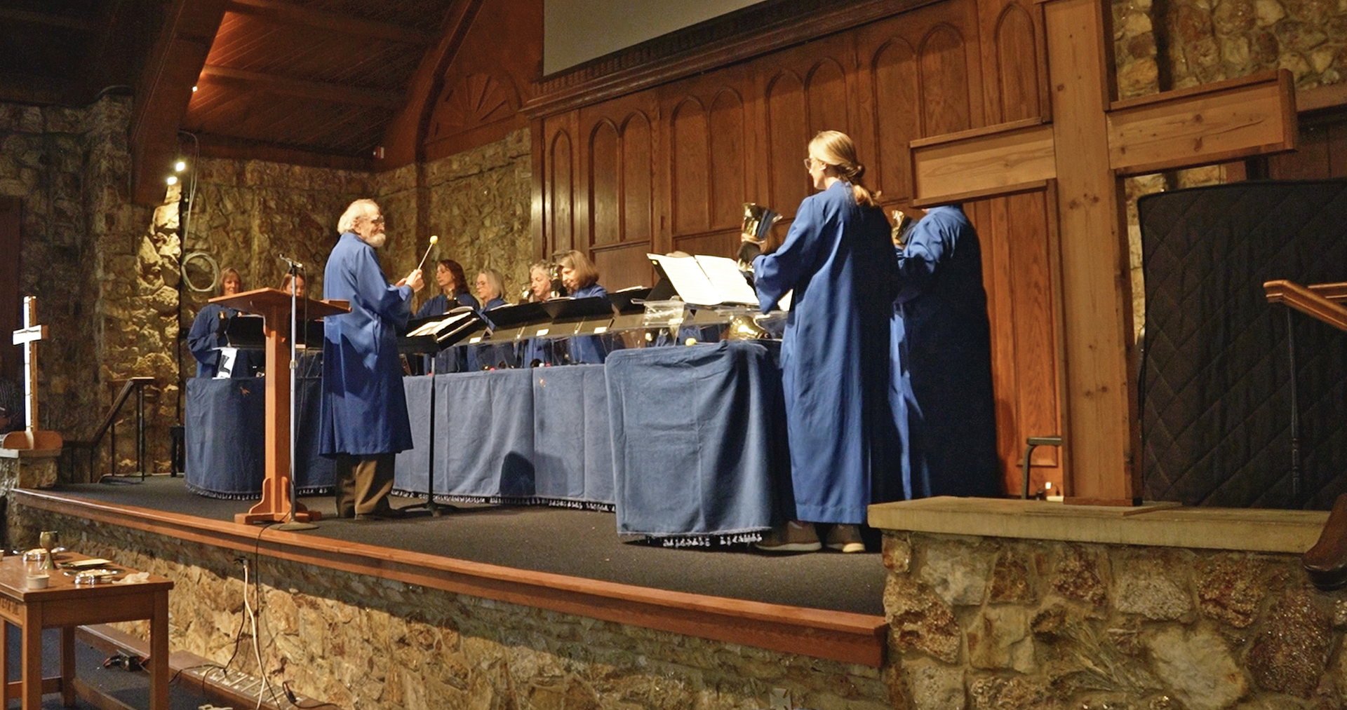 A group of people wearing blue robes playing handbells in a stone building with wood accents.
