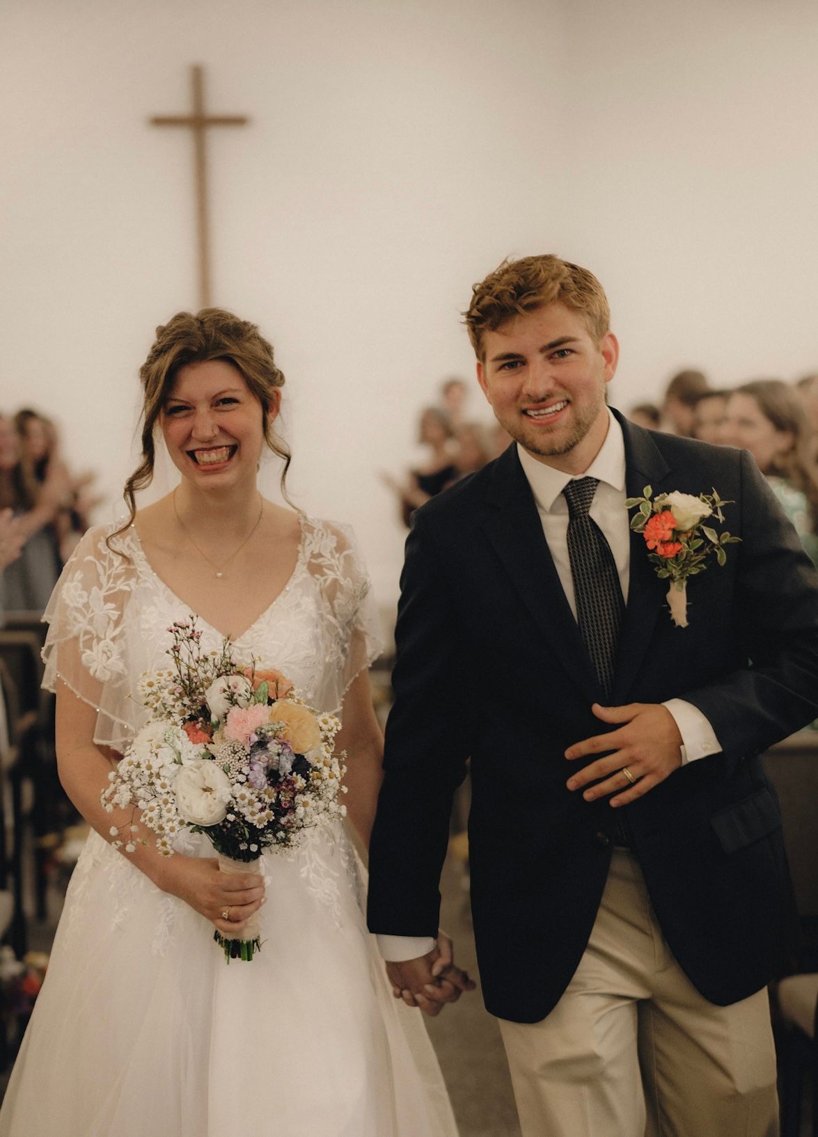 Bride and groom walking down the aisle, holding bouquets, with a cross in the background