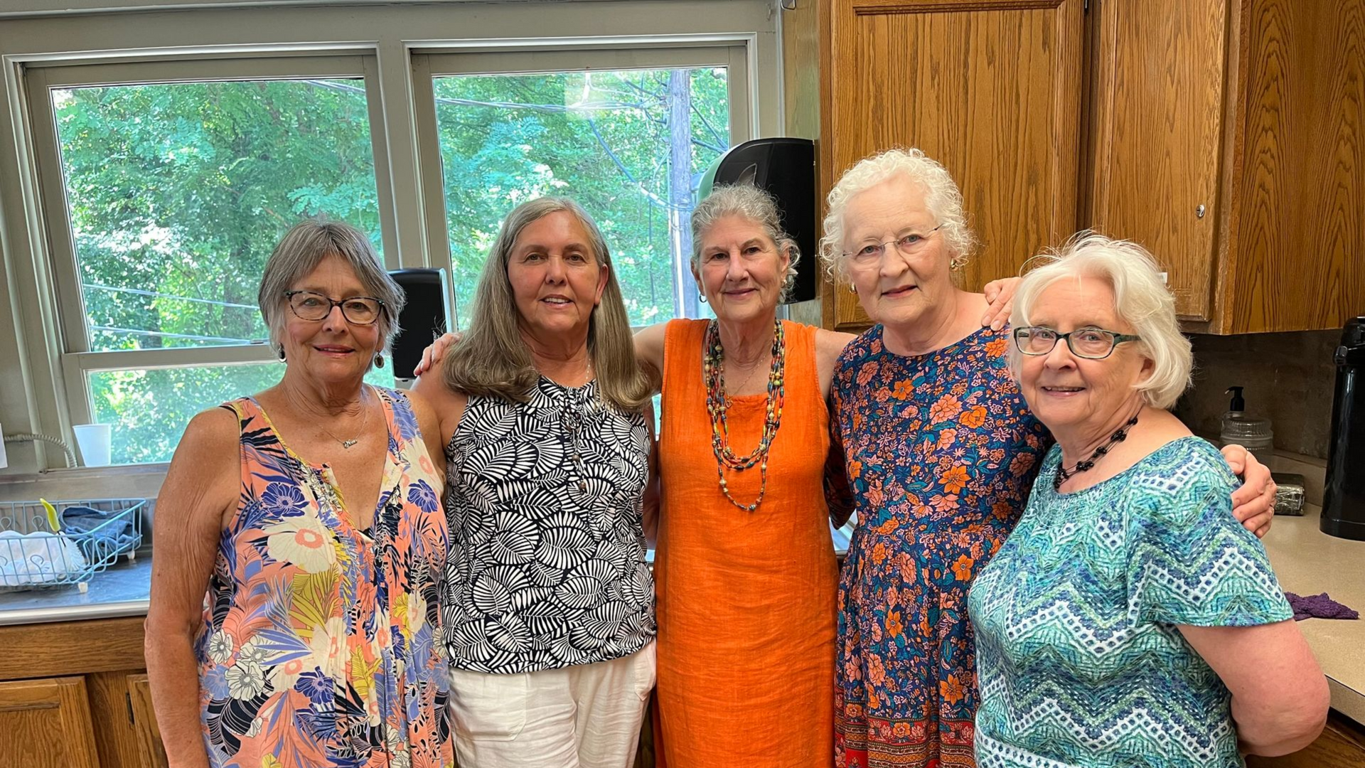 Five women stand together smiling in a kitchen with a window, wooden cabinets, and colorful tops.