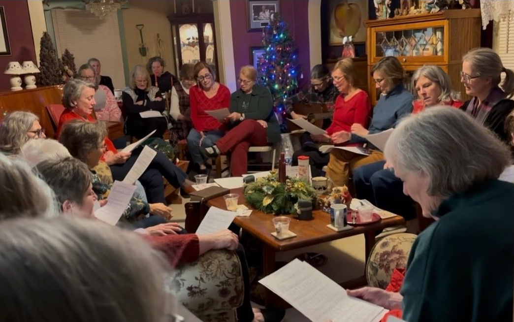 A large group of women gathered in a living room holding sheets of music to sing.
