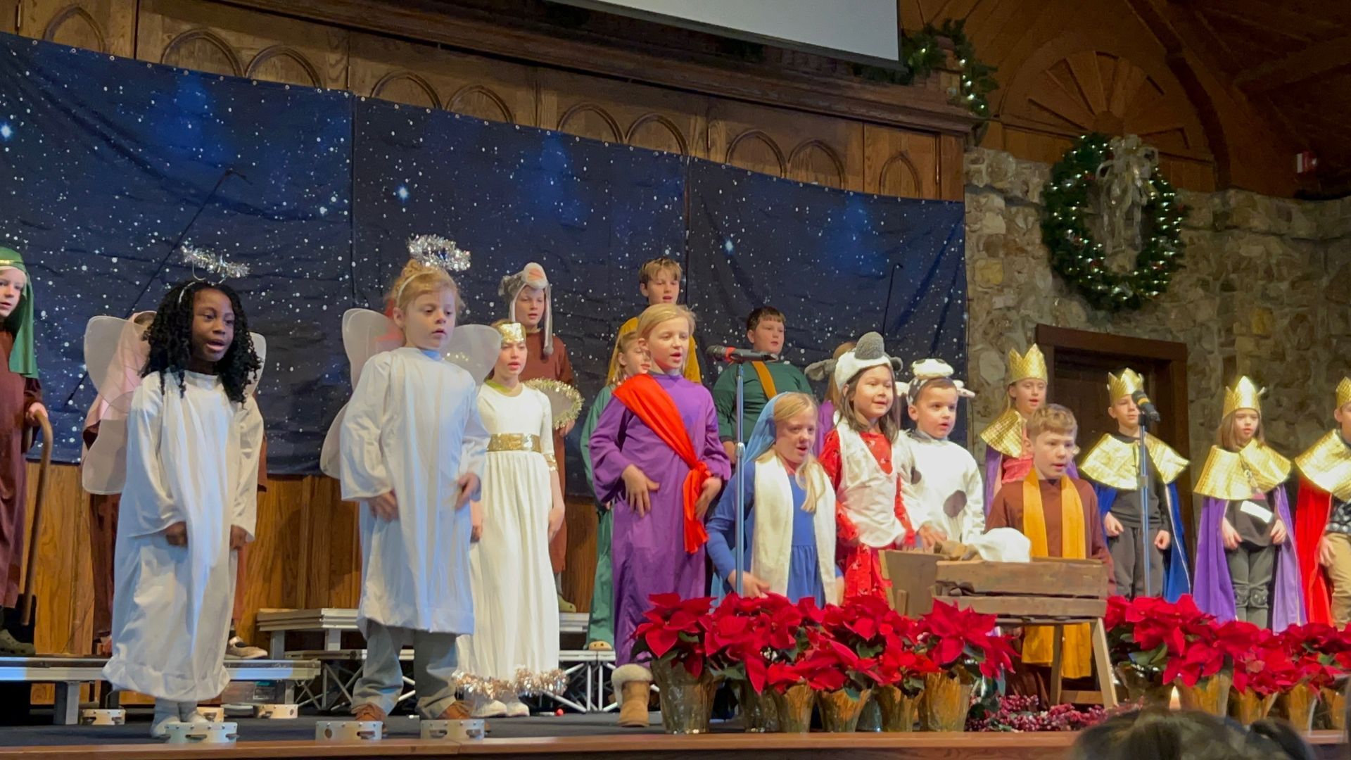 Children in Christmas Bible costumes sing on a stage with poinsettias and holiday decorations.