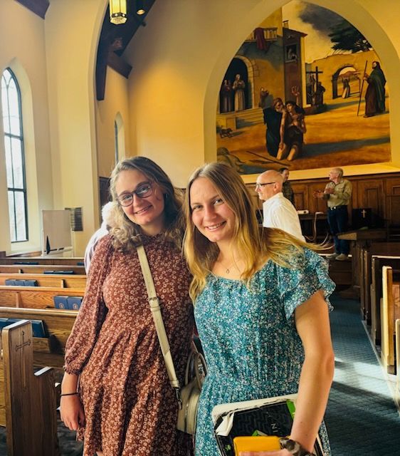 Two smiling people pose in a church pew aisle beneath a mural and tall windows.
