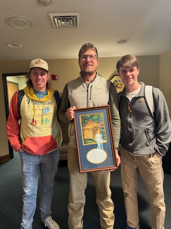 Three people standing indoors; center person holds a framed award or artwork.