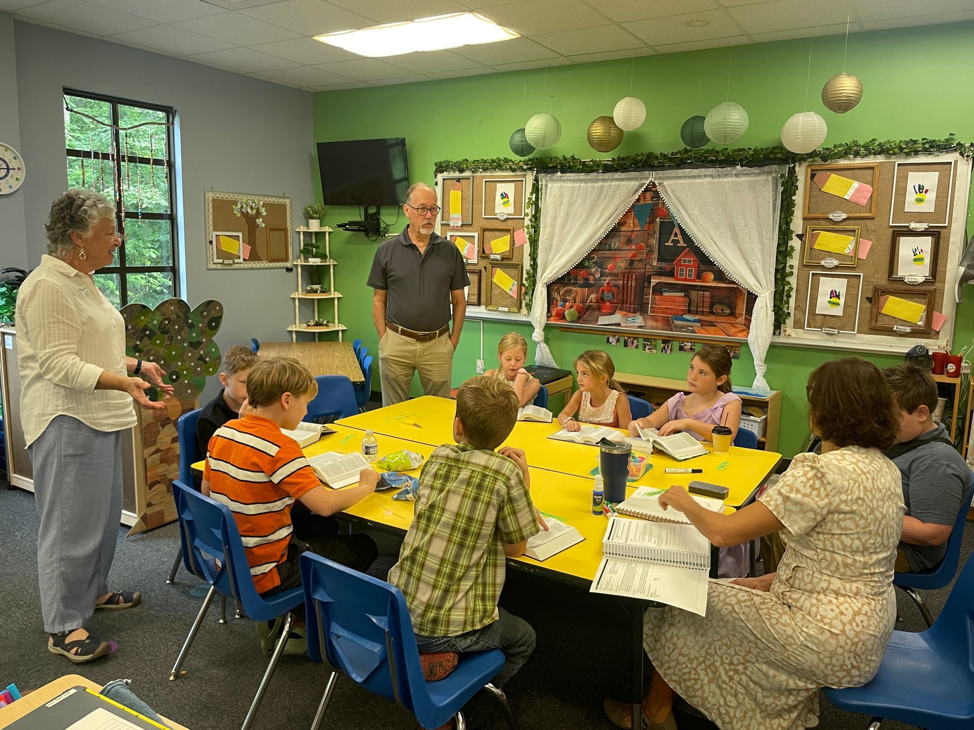 Children seated around a yellow table in a classroom, with an adult standing nearby and wall art in the background
