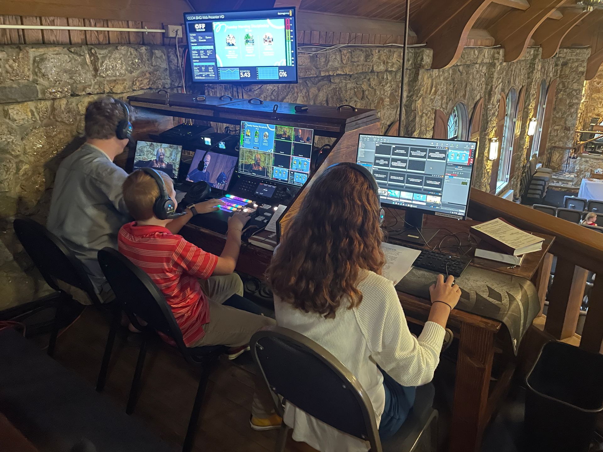 Three people sit in a row at a wooden desk with computer monitors in a rustic, stone-walled room.