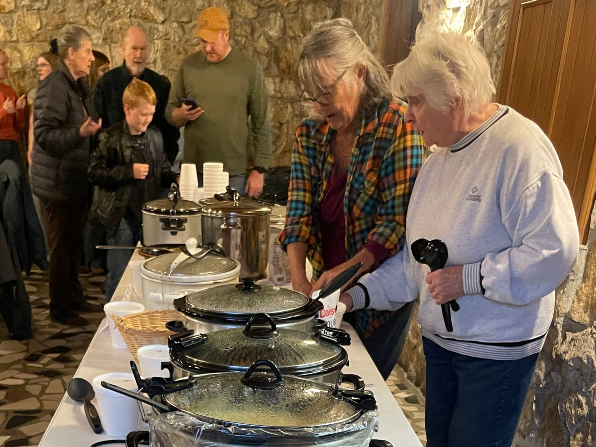 People serving food from a buffet line in a busy indoor gathering.