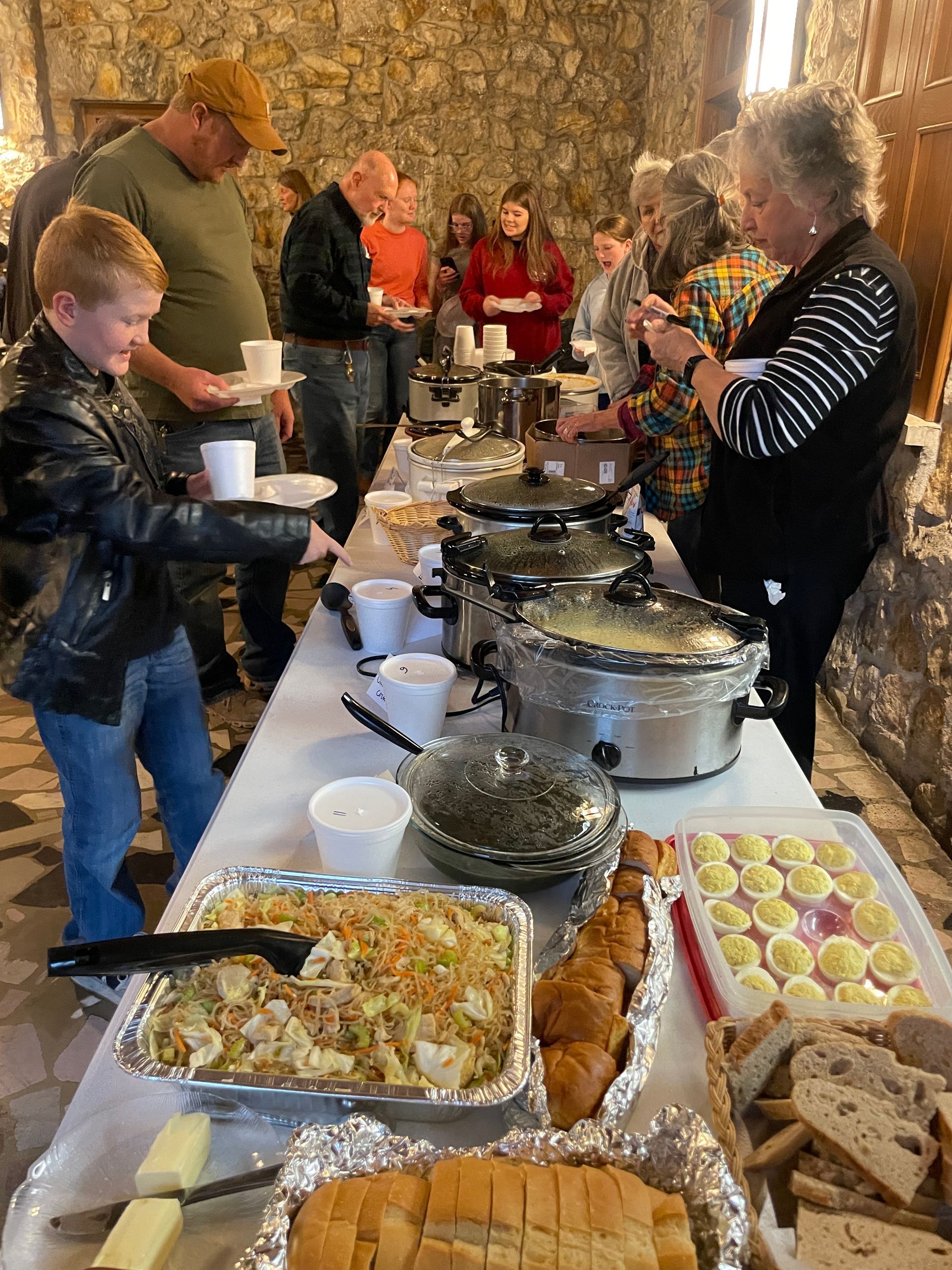 Buffet table with covered dishes, desserts, and bread as people serve themselves in a stone-walled room.