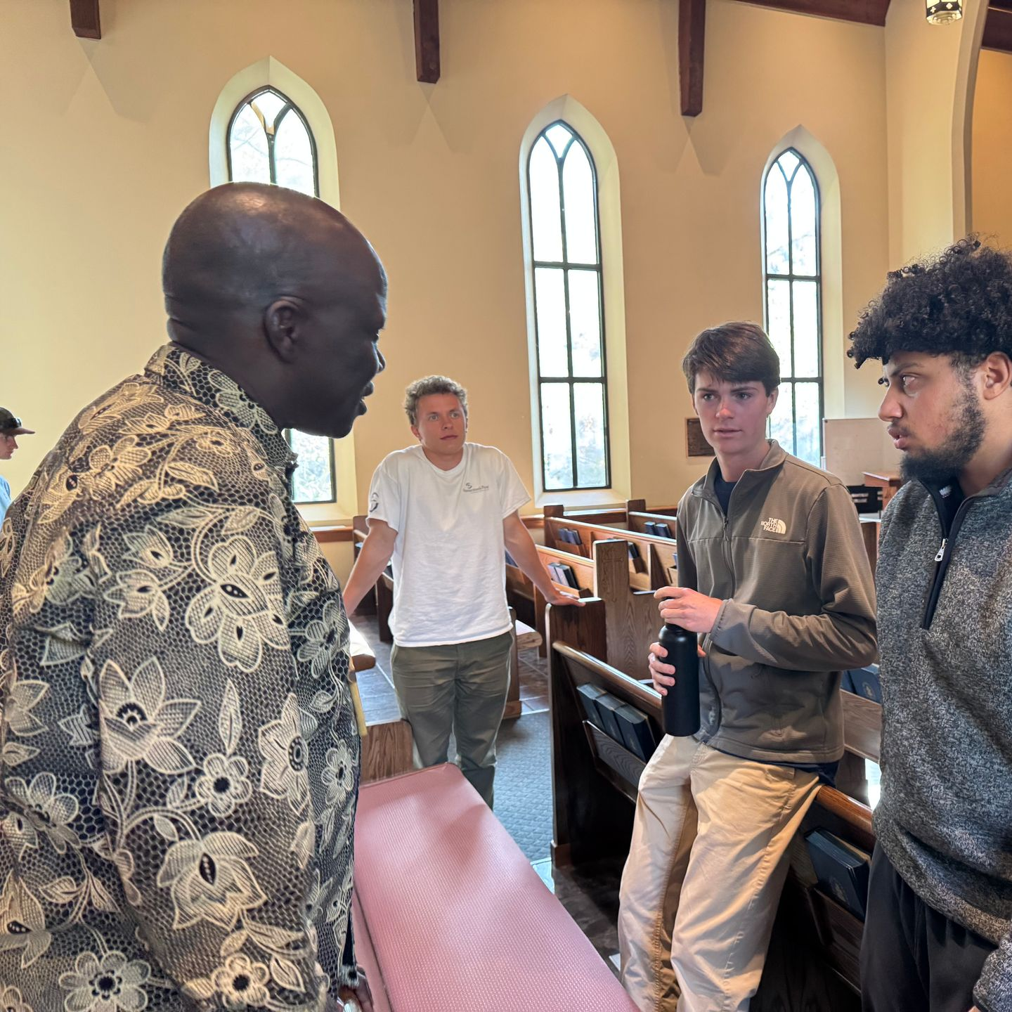 People conversing inside a church, with pews and tall arched windows in the background.