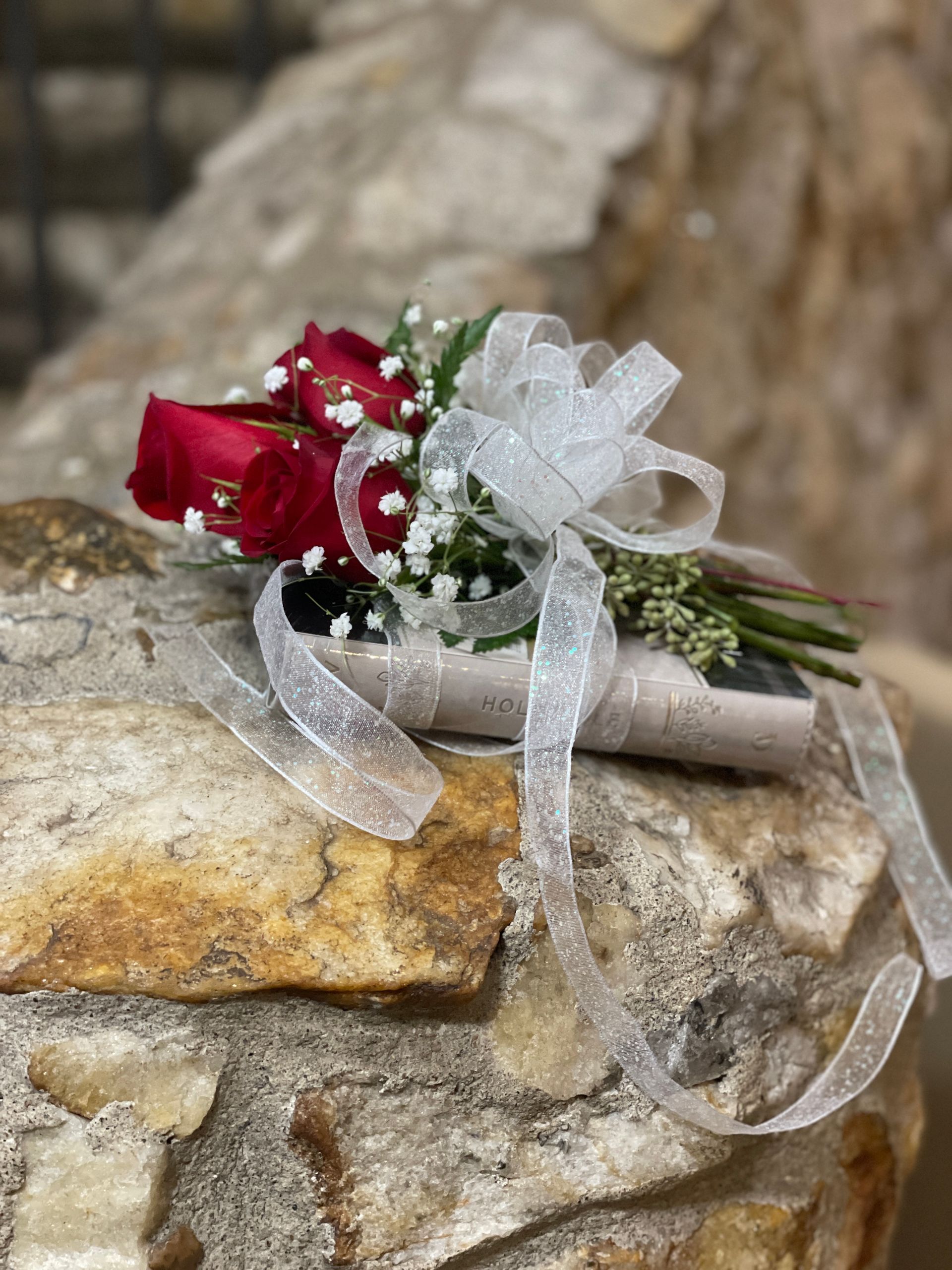 Red rose buds tied in white ribbon onto a Bible resting on stonework.