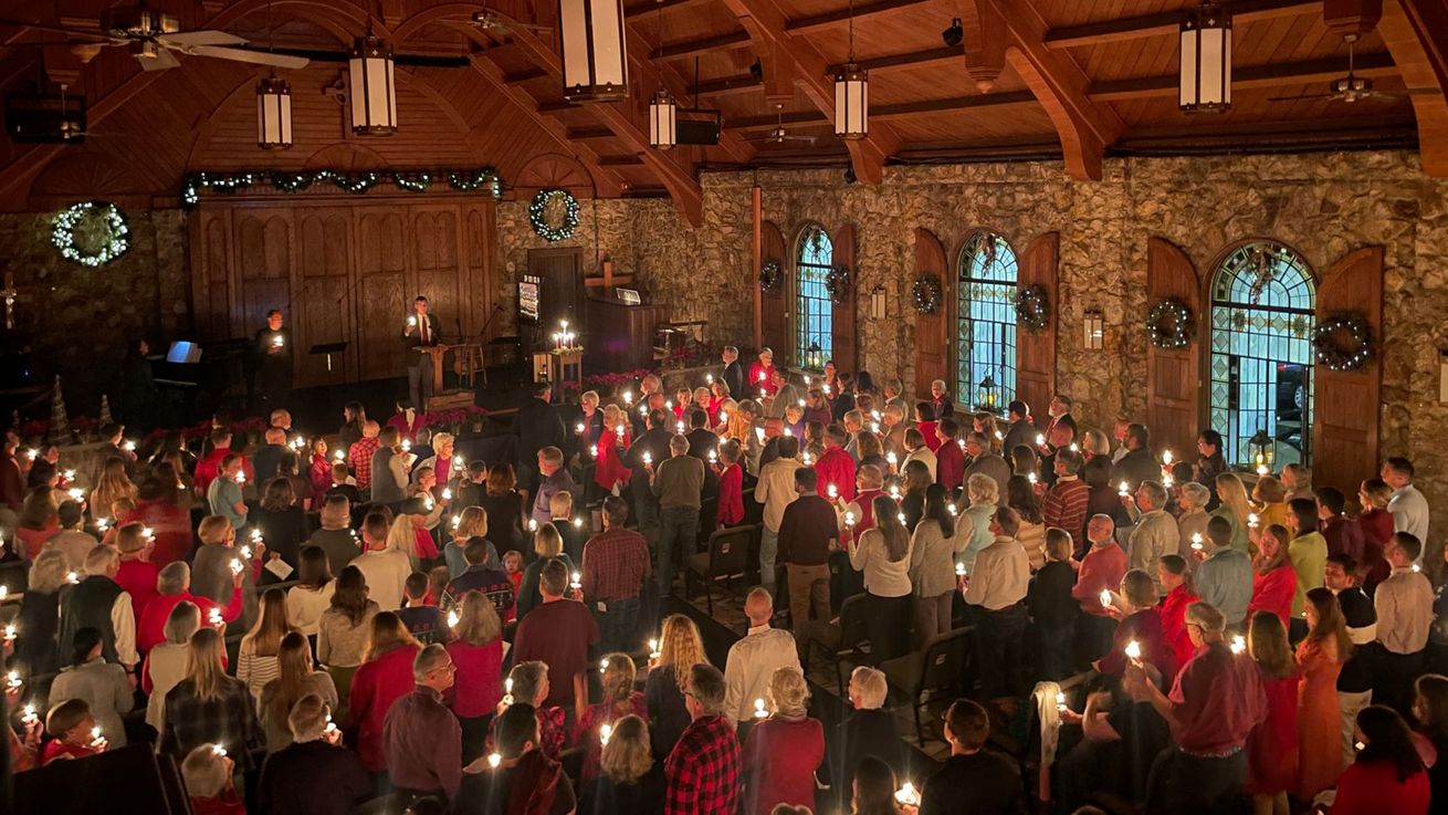 Aerial view of people holding candles in a stone chapel with lighted Christmas greenery.