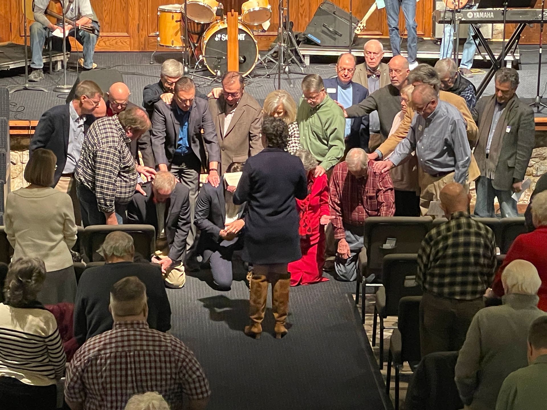 A group of people standing on a stage at an indoor event, with a speaker addressing them from the foreground.