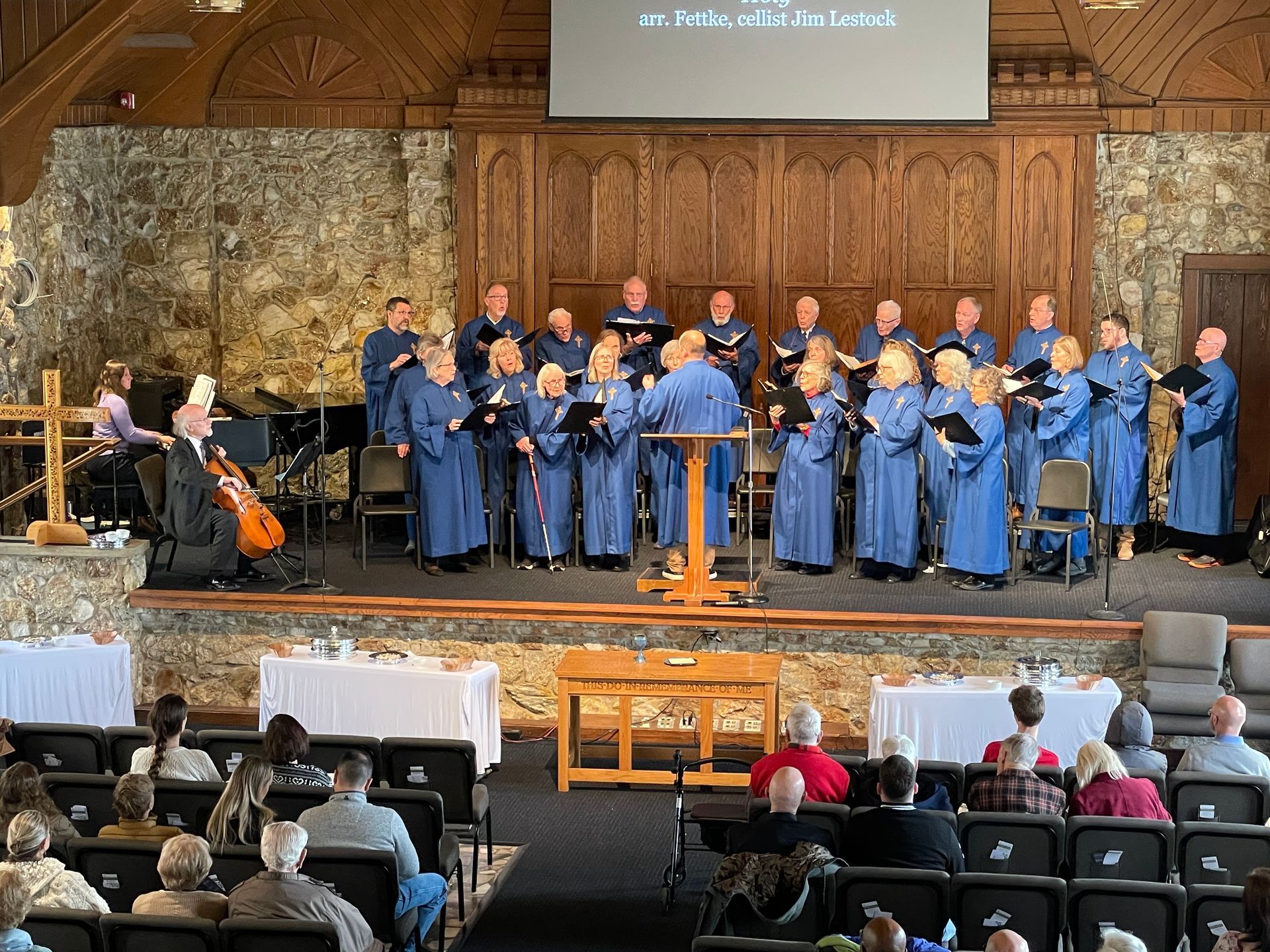 Choir in blue robes performing on a church stage with cello and piano and audience seated below.