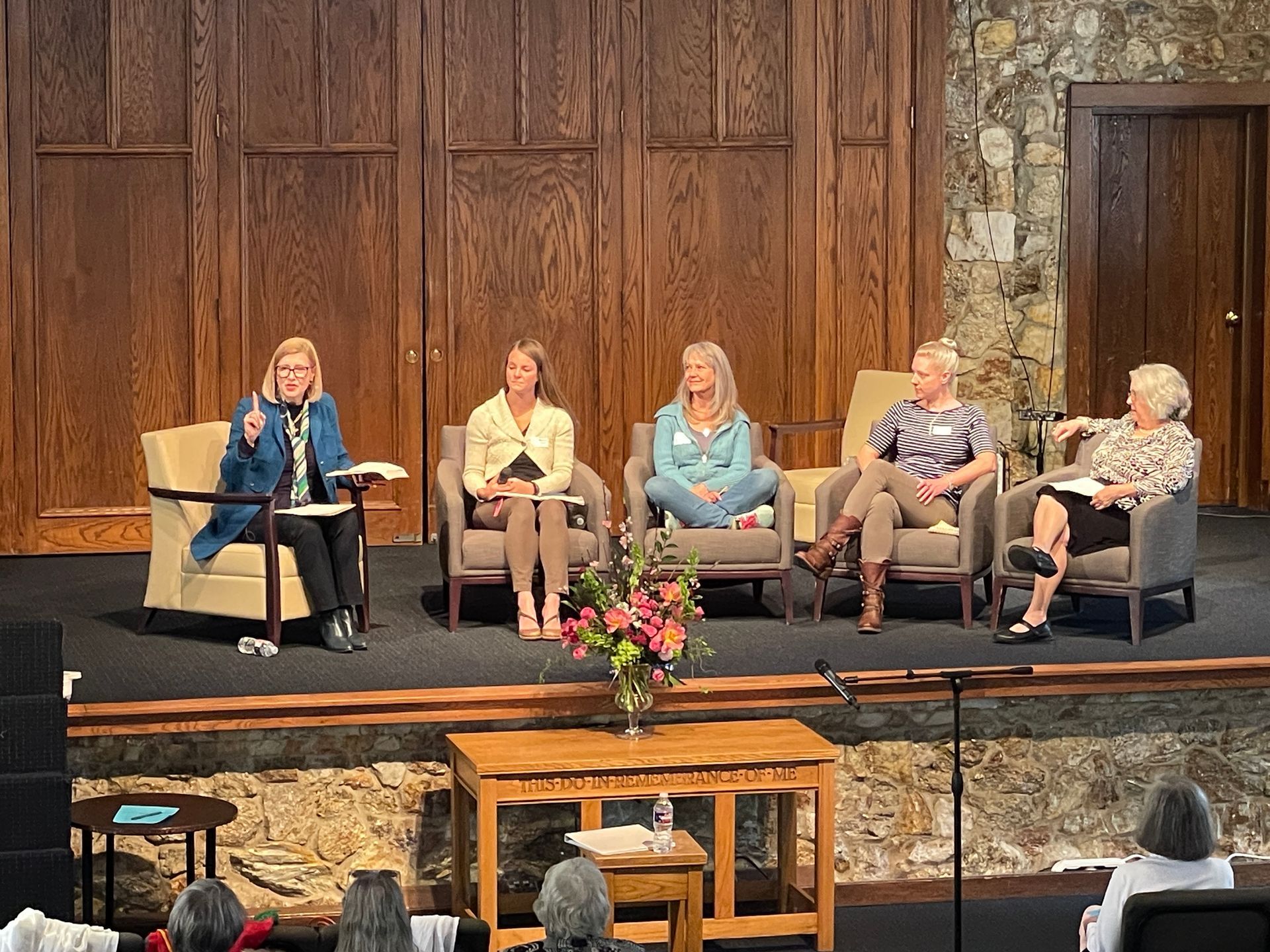 Panel discussion on stage with five speakers seated before a wooden backdrop and flower arrangement.