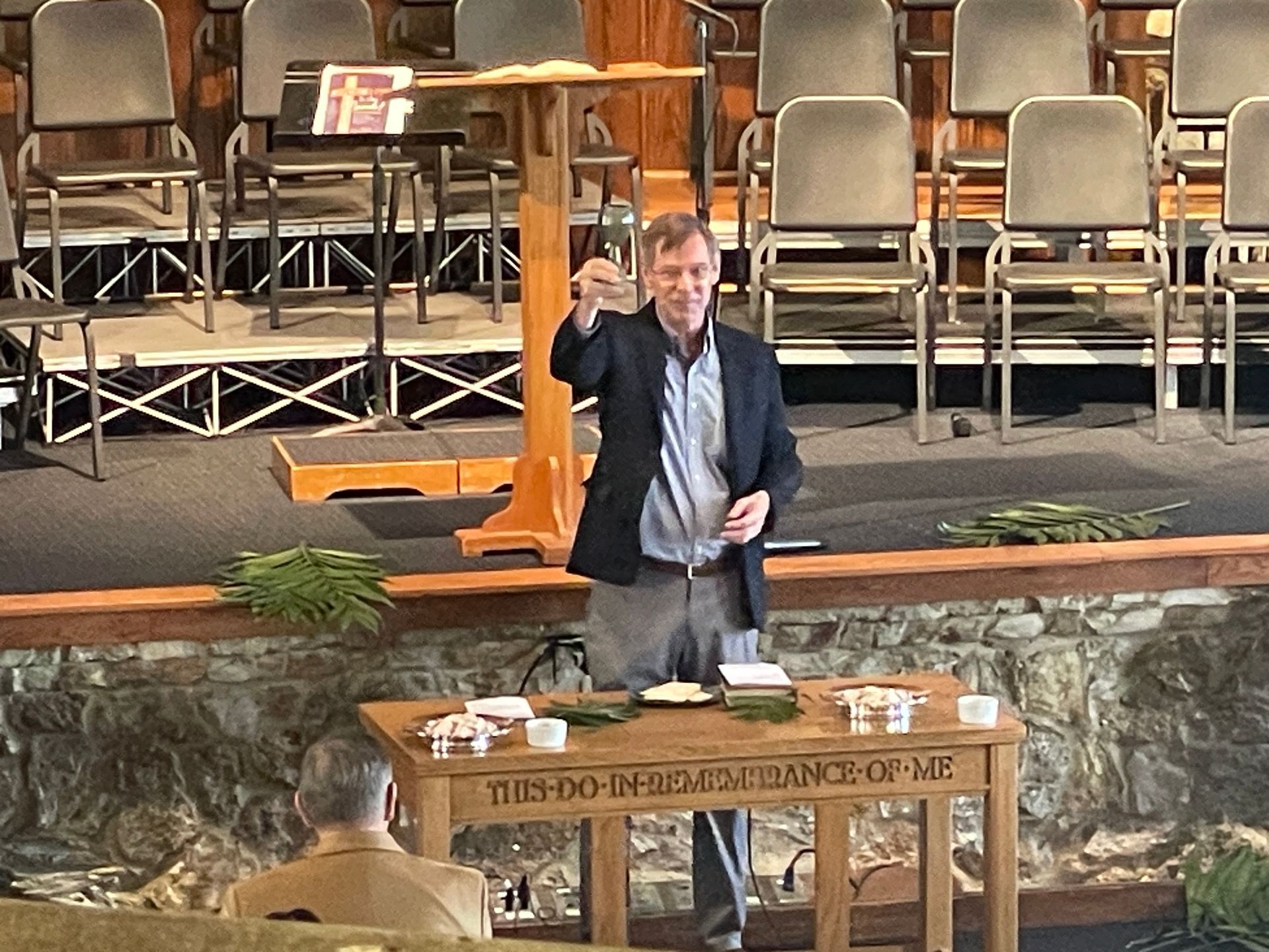 The pastor stands behind a wooden communion table in a church, holding up a chalice.