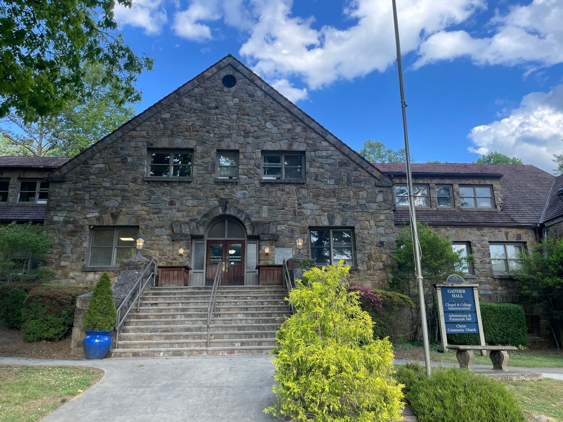 Stone building with central steps and arched doorway, under a blue sky with trees and a sign outside